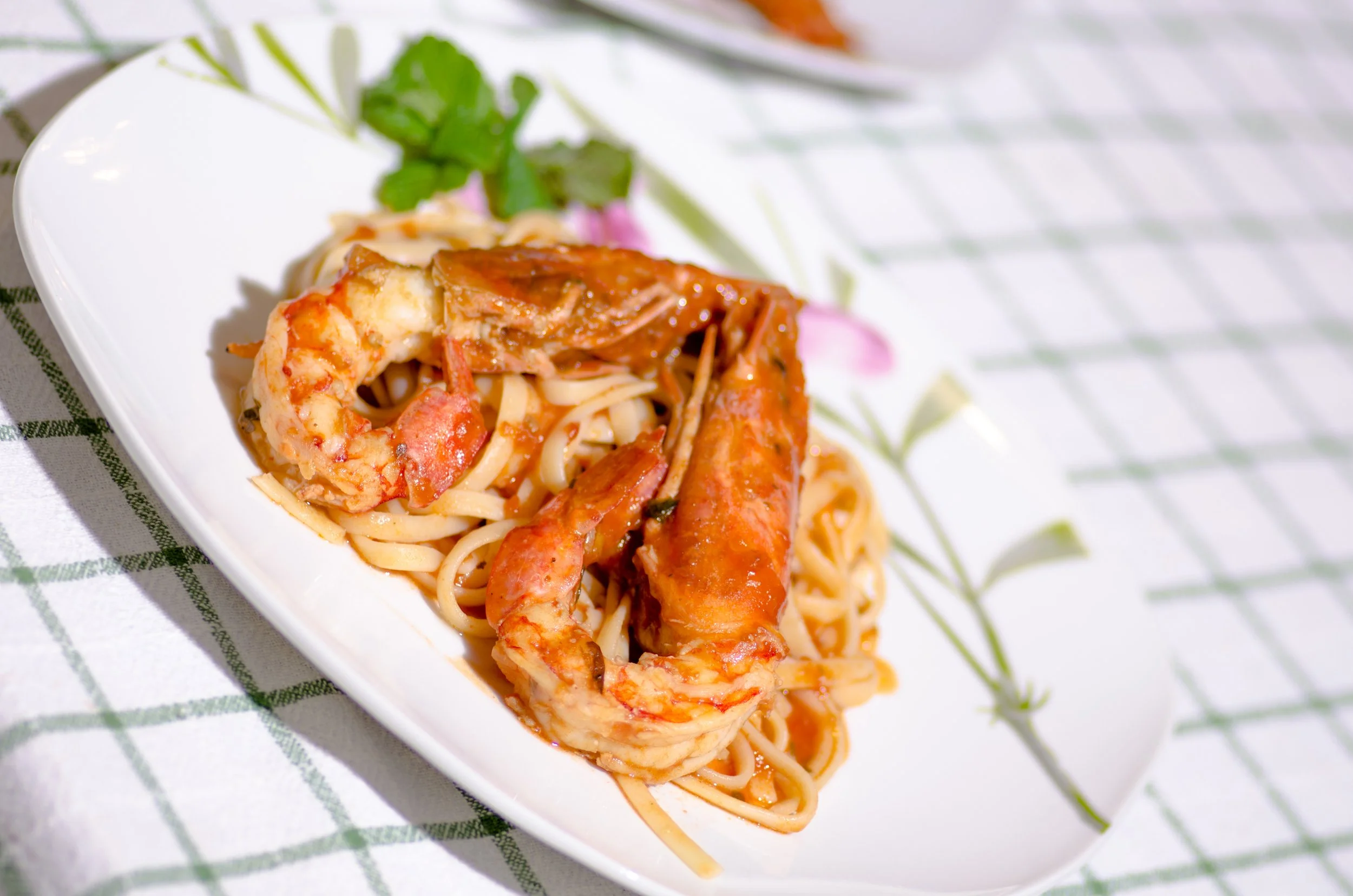 Close-up of a bowl of pasta with shrimp and tomato sauce garnished with herbs, placed on a white checkered cloth.