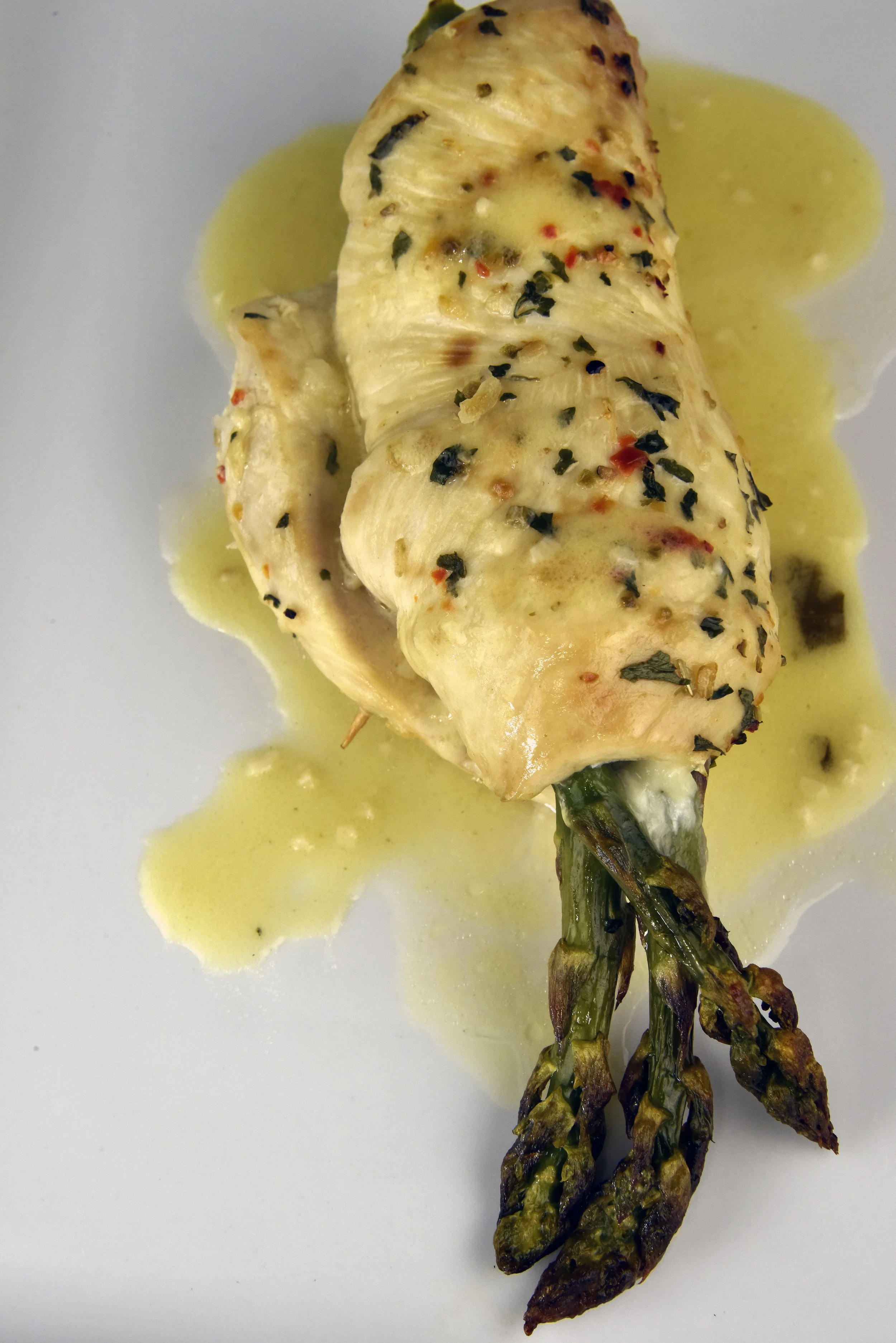 A roasted asparagus spear topped with melted cheese, herbs, and red pepper flakes, served with a creamy yellow sauce.