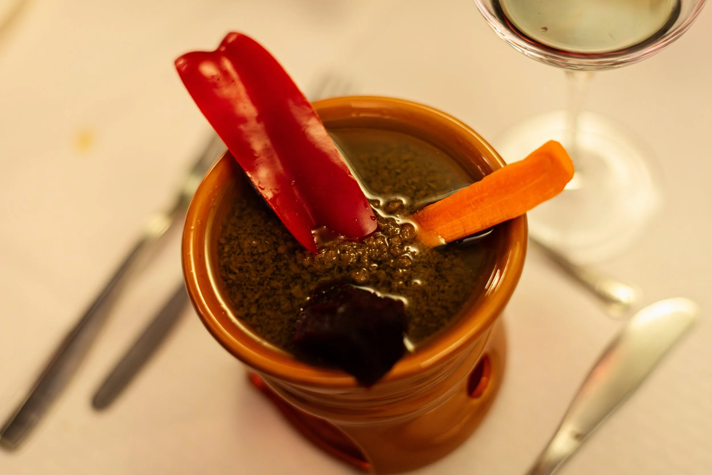 Close-up of a drink with a red chili pepper and an orange carrot slice garnishing the rim of a glass, with a partially visible glass of white wine in the background.