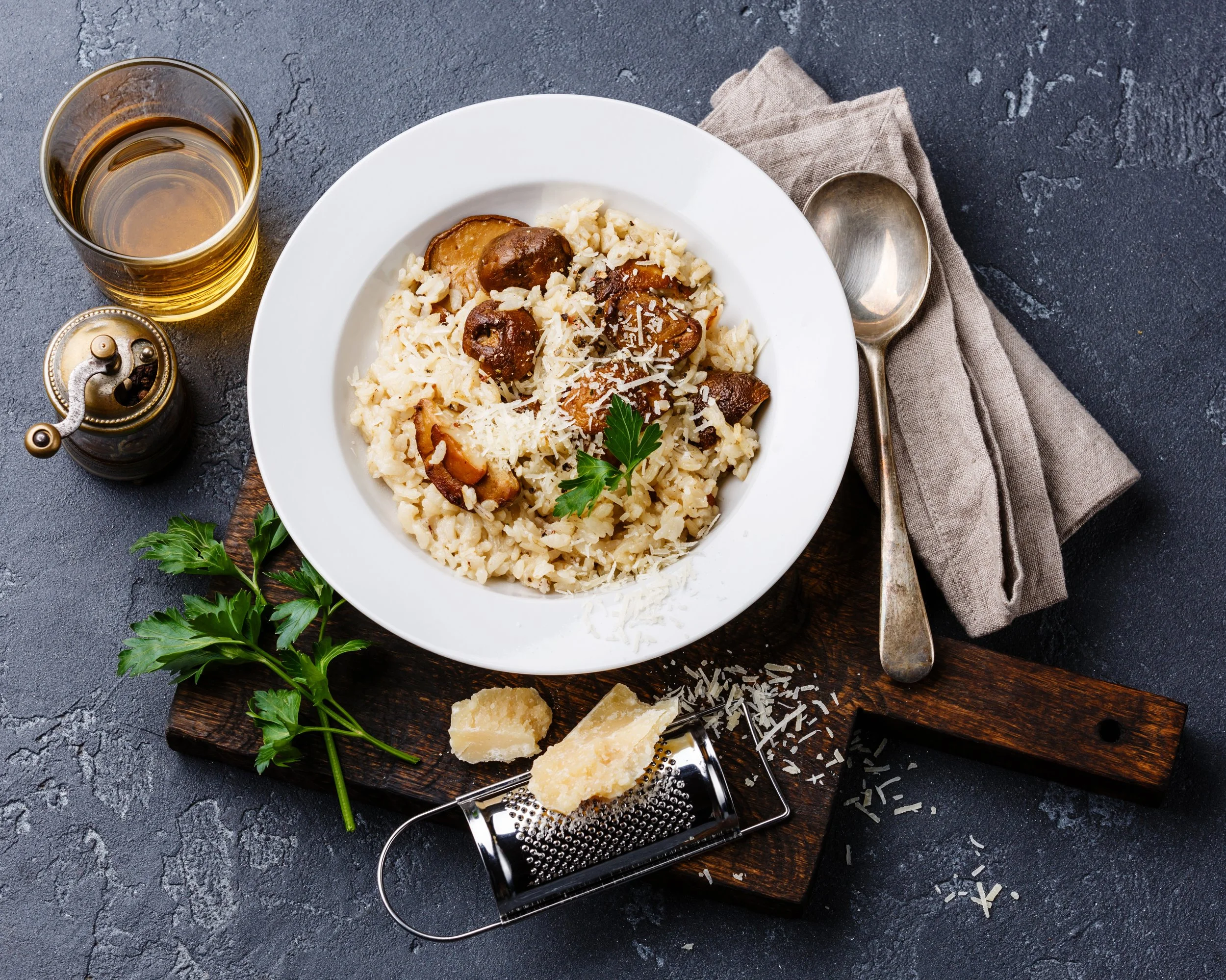A plate of creamy risotto with mushrooms and grated cheese, garnished with a sprig of parsley. Accompanying are a glass of white wine, a chunk of Parmesan cheese, and a cheese grater, all placed on a rustic wooden board with a gray napkin.