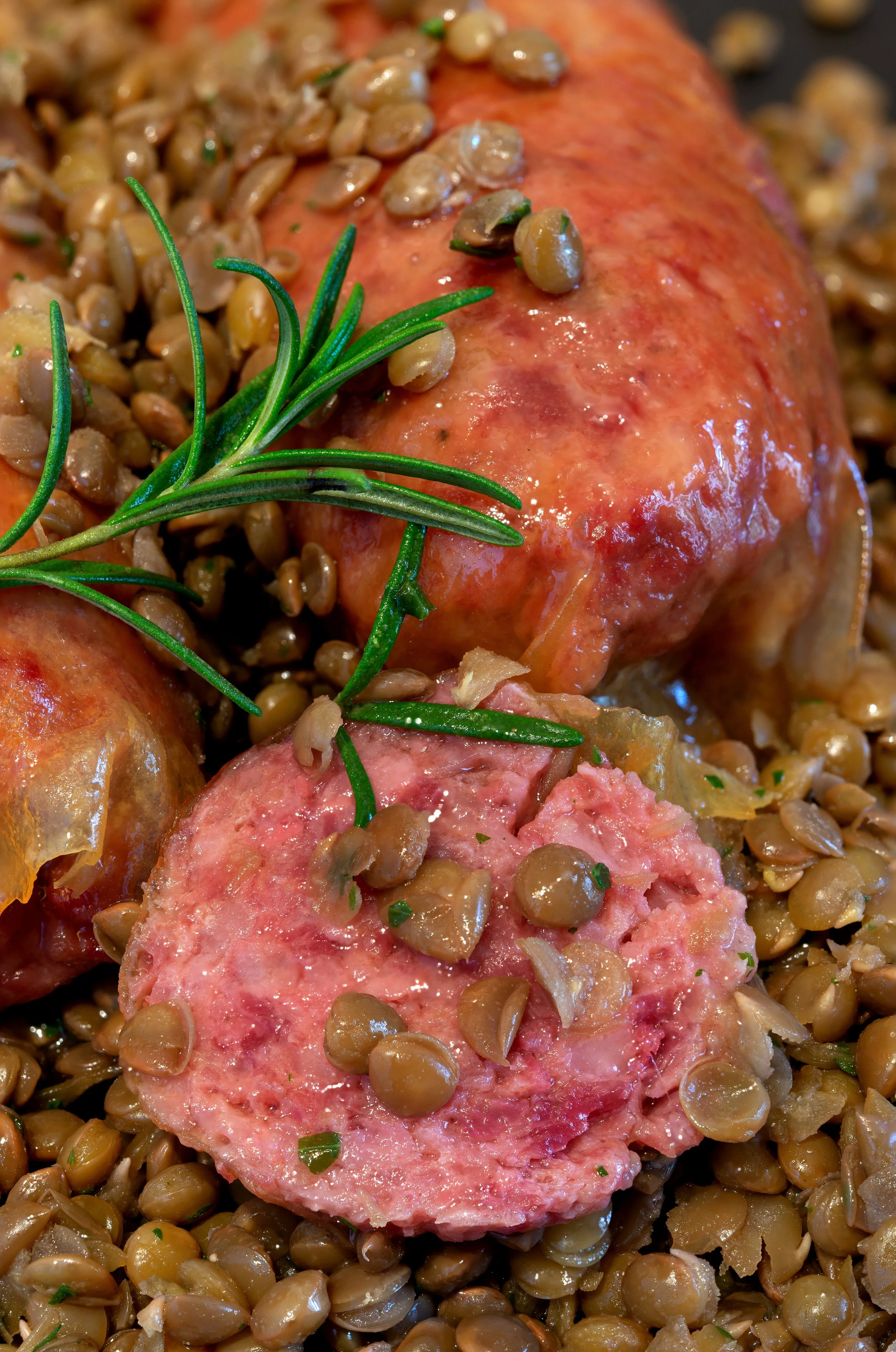 Cooked lentils garnished with a sprig of rosemary, with sliced sausage and salt in the background.