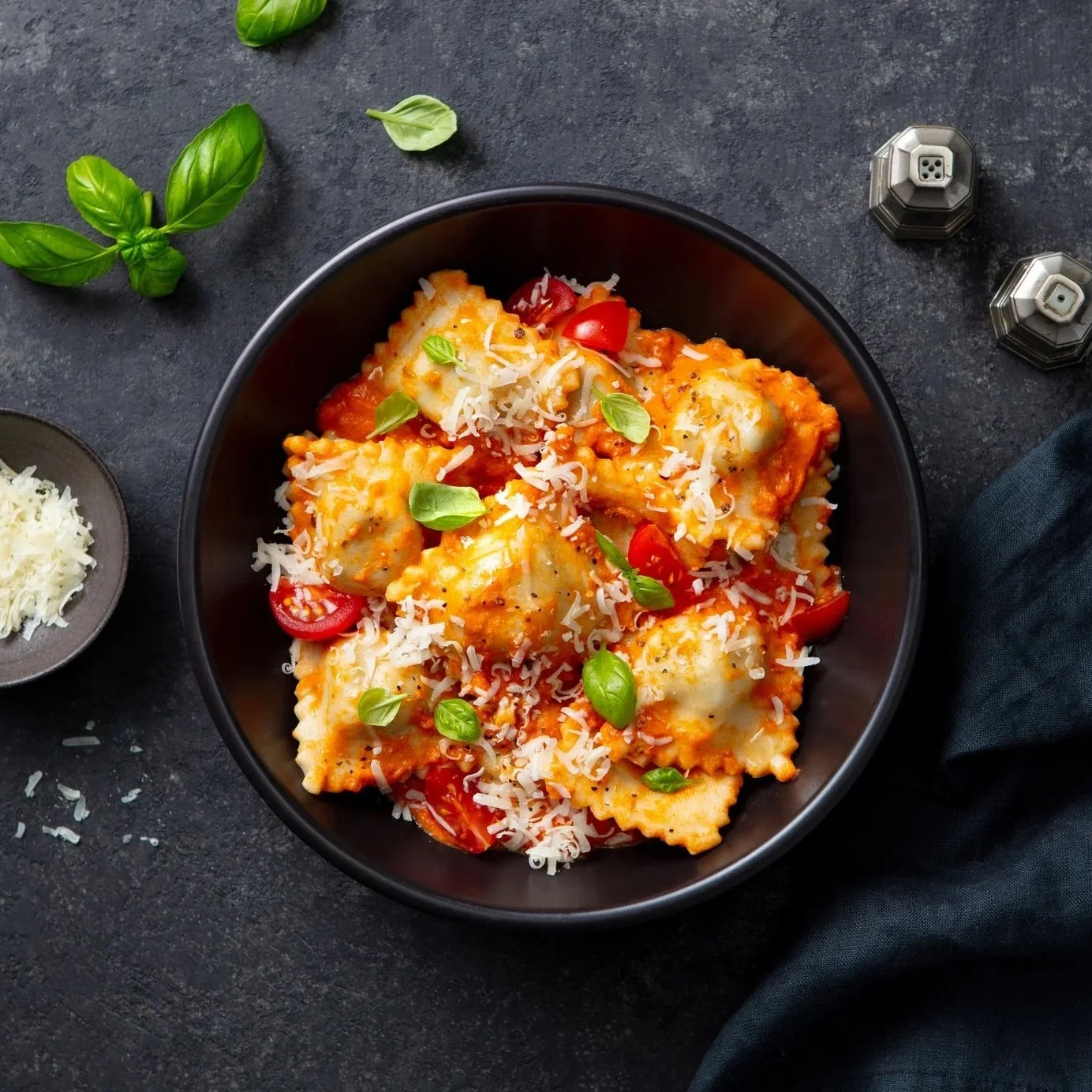 Bowl of ravioli pasta with tomato sauce, cherry tomatoes, shredded cheese, and fresh basil leaves on top, placed on a dark surface with basil sprigs nearby.