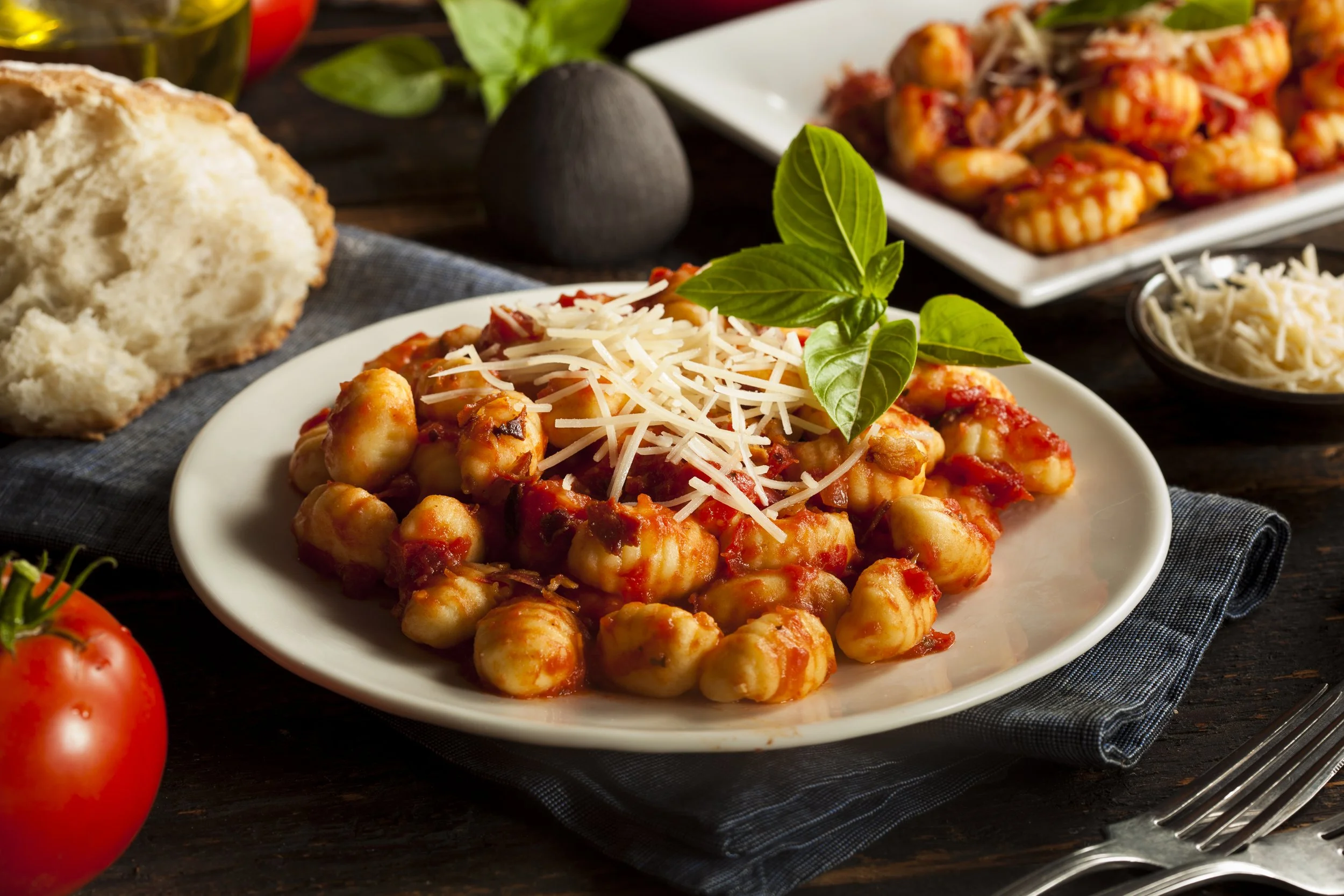 Plate of gnocchi with tomato sauce, grated cheese, and basil garnished with basil leaves, on a dark wooden table with tomatoes and bread nearby.