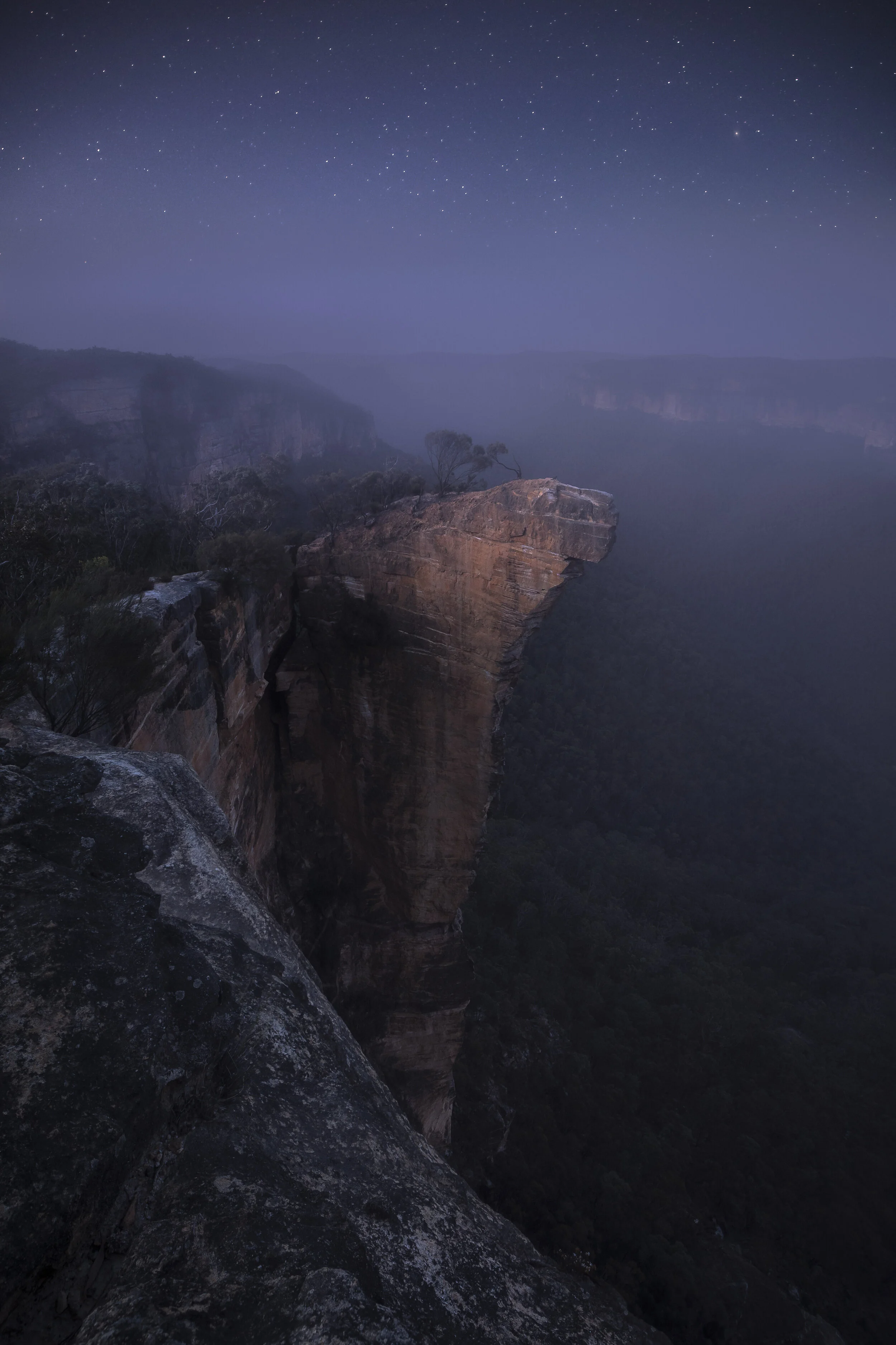 Stars twinkling in the night sky above Hanging Rock in the Blue Mountains, Australia, with smoke drifting over cliffs and forest