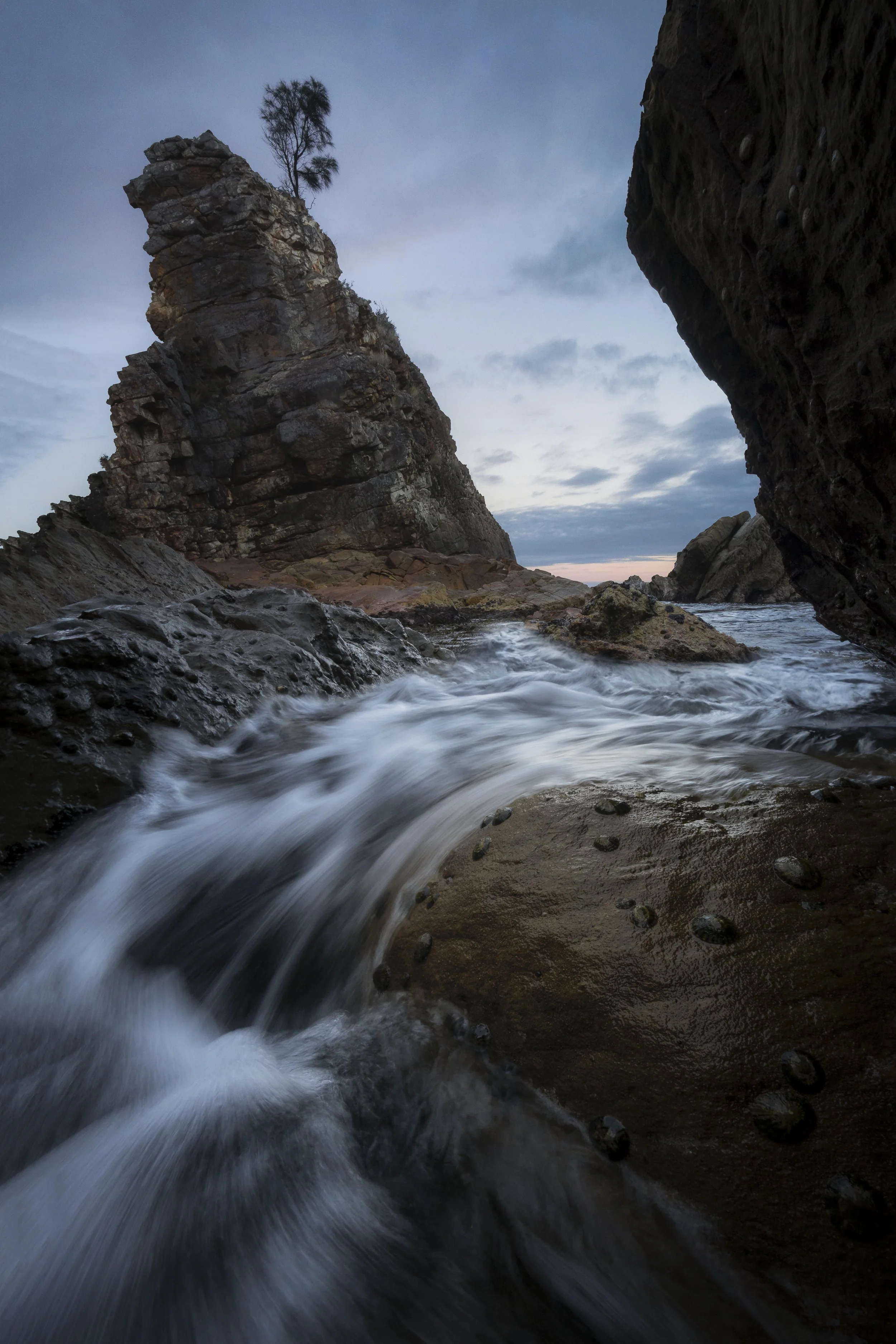 Long exposure of waves flowing around over rocks with a sharp rock in the background