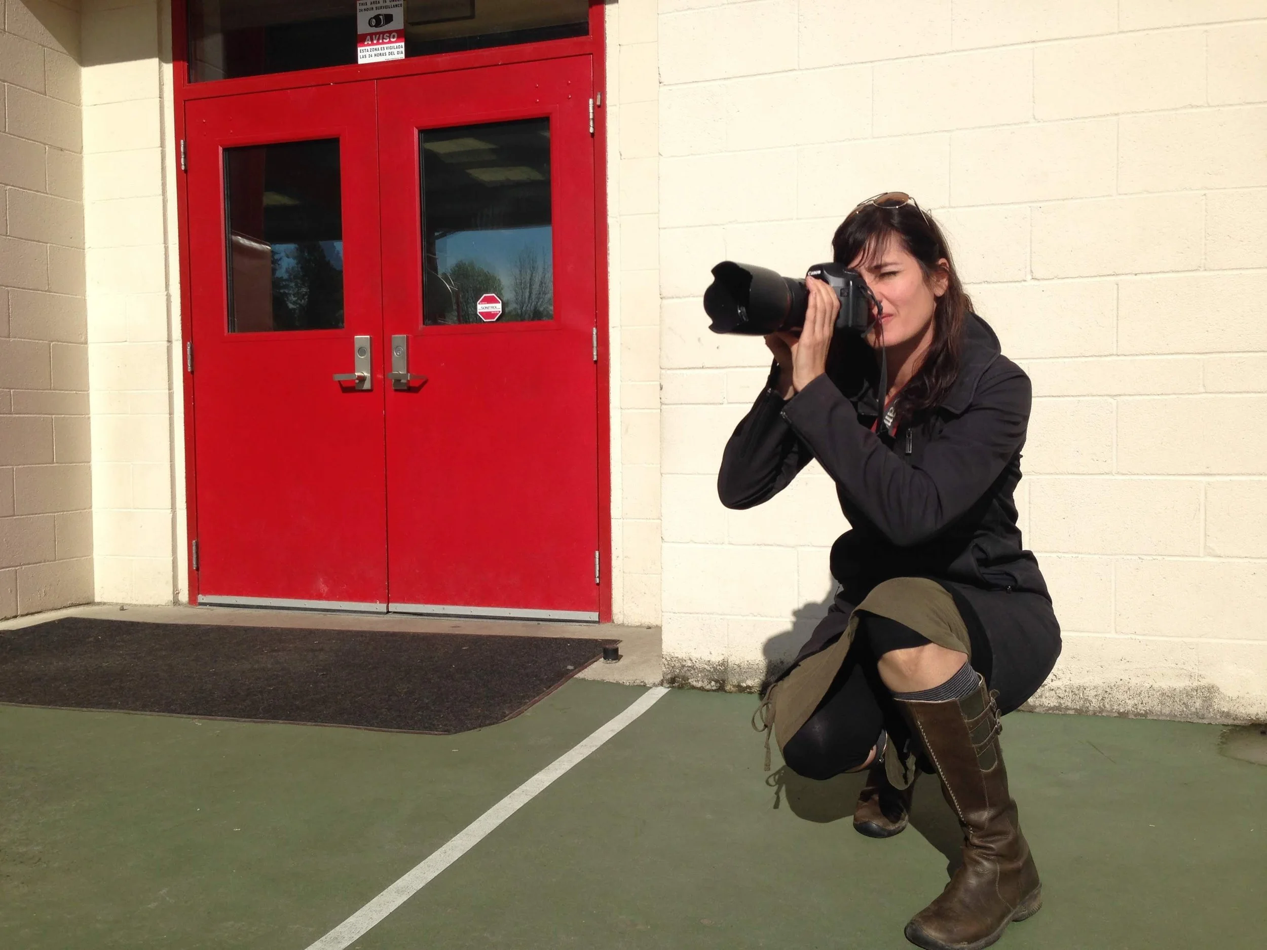 Person dressed as a cowboy, crouching with a camera, positioned outdoors near a red double door with glass panels.