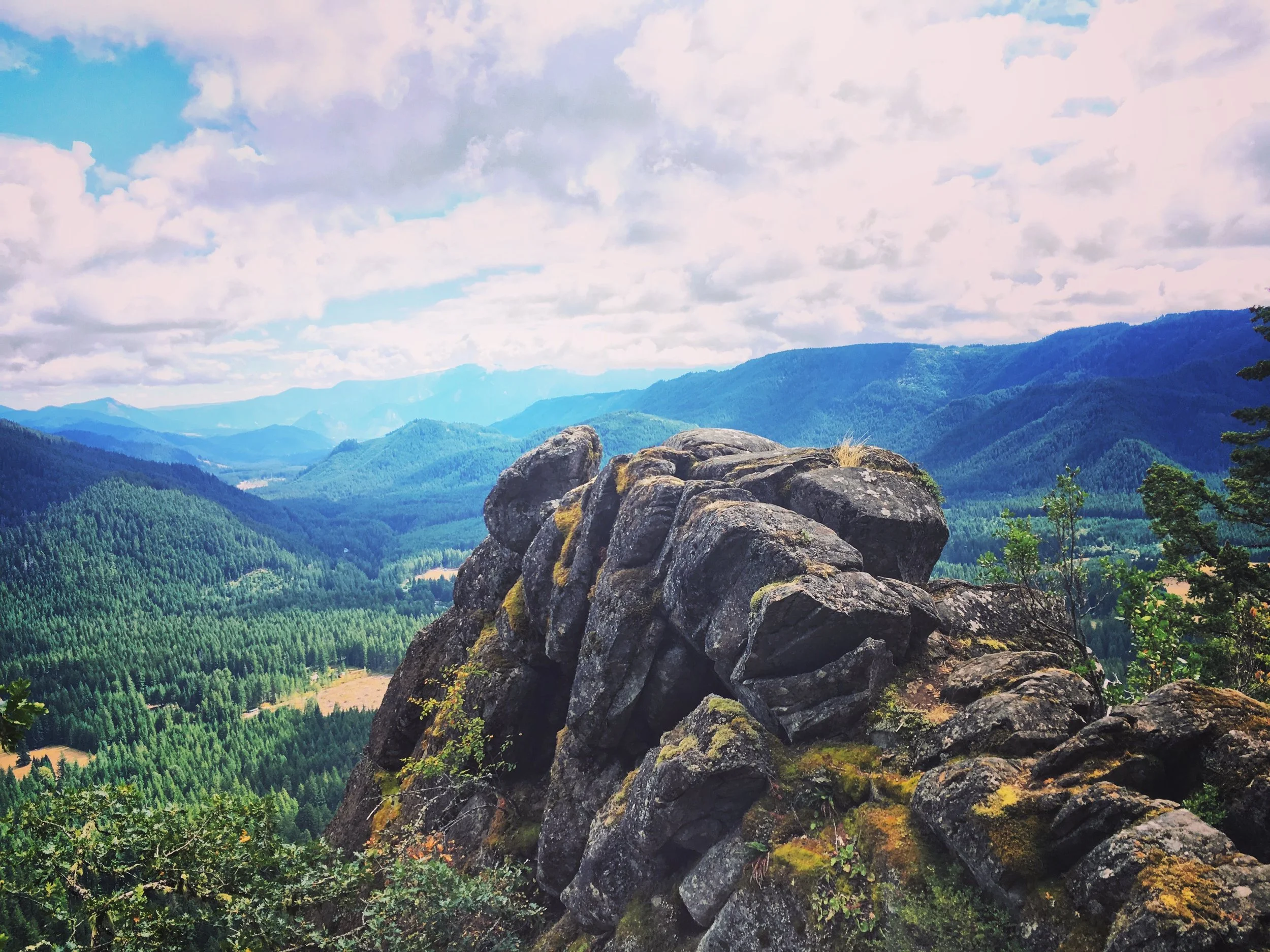 A scenic view of a mountain range covered in dense green forests under a partly cloudy sky, with large rocks in the foreground.