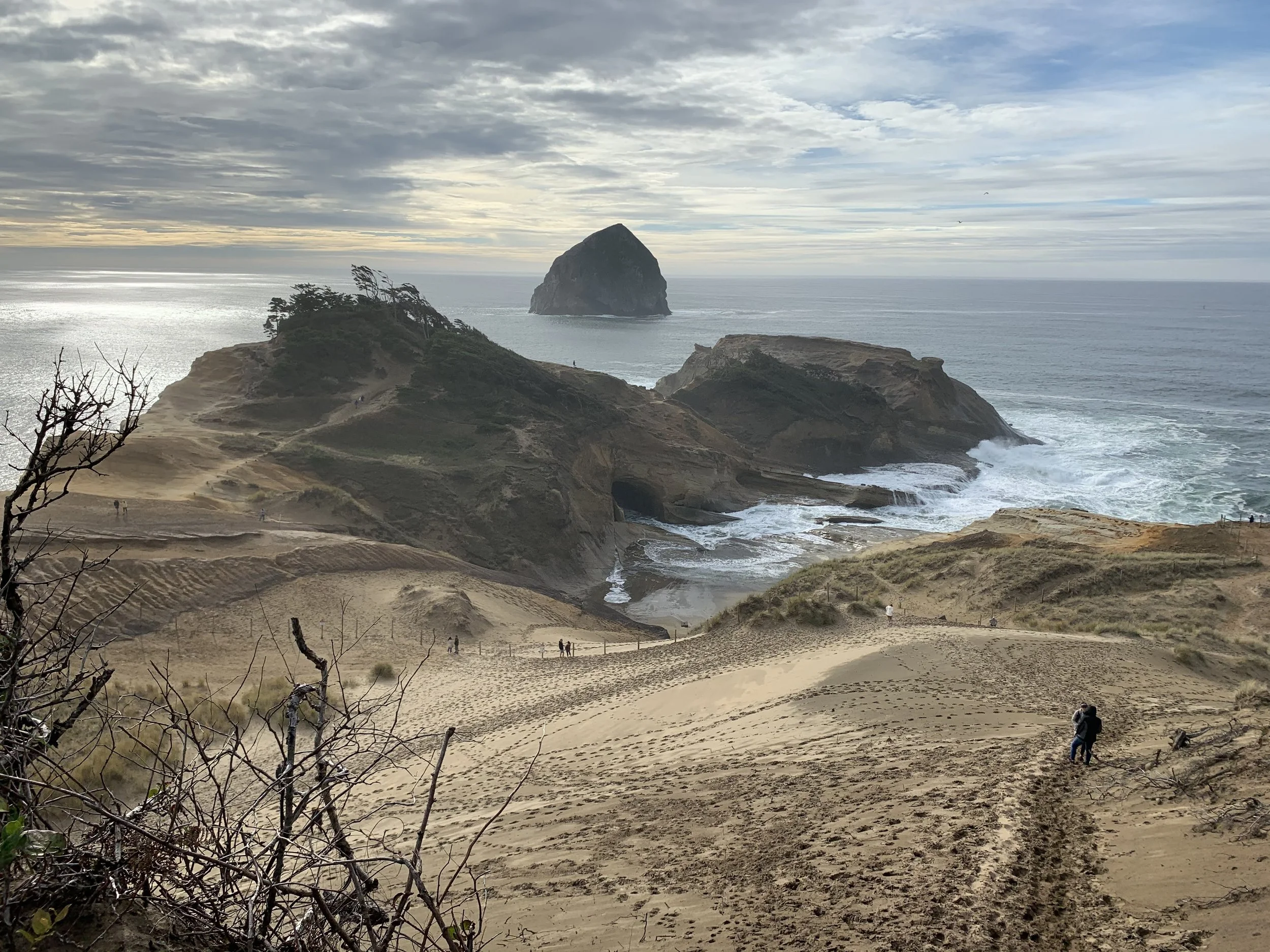 View of a sandy beach with footprints, overlooking cliffs and the ocean with a large rock formation in the water under a cloudy sky.