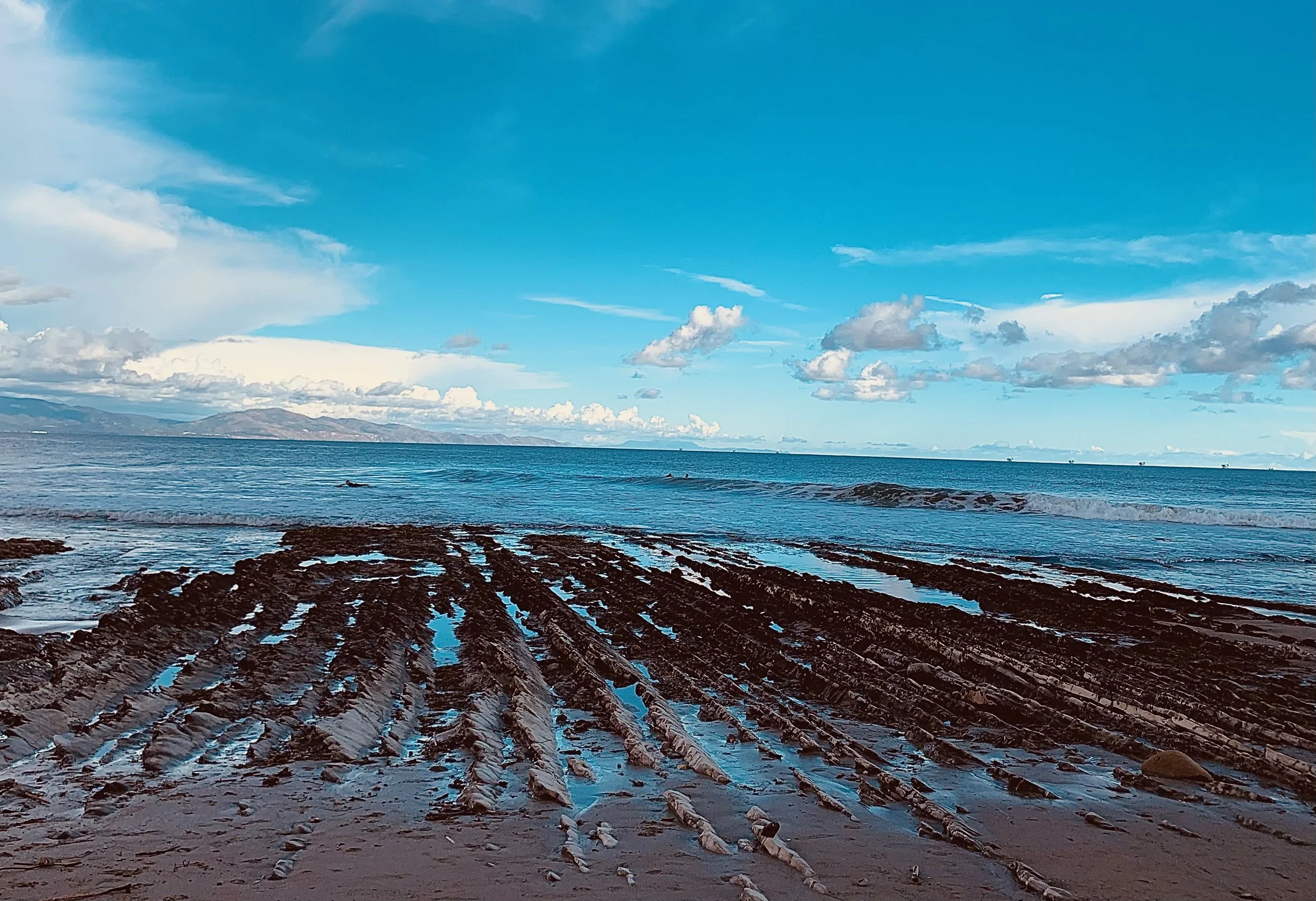 Coastal scene with rocky shore, calm water, and a bright blue sky with scattered clouds.