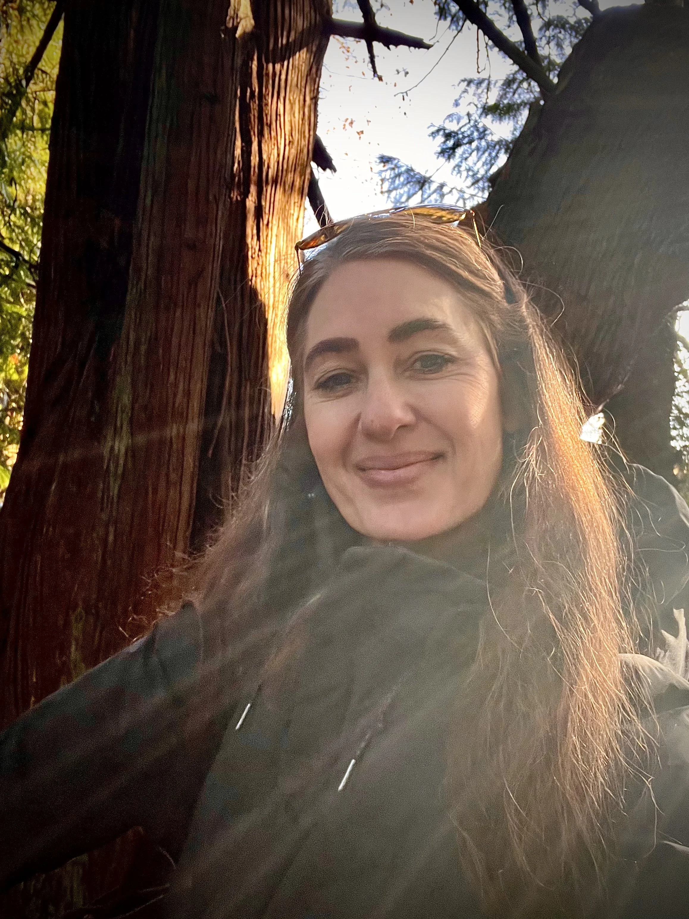 A woman with long brown hair taking a selfie outdoors, with trees and sunlight visible behind her.