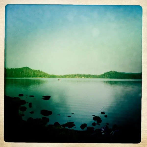 A calm lake with a rocky shoreline and distant tree-covered hills under a partly cloudy sky.