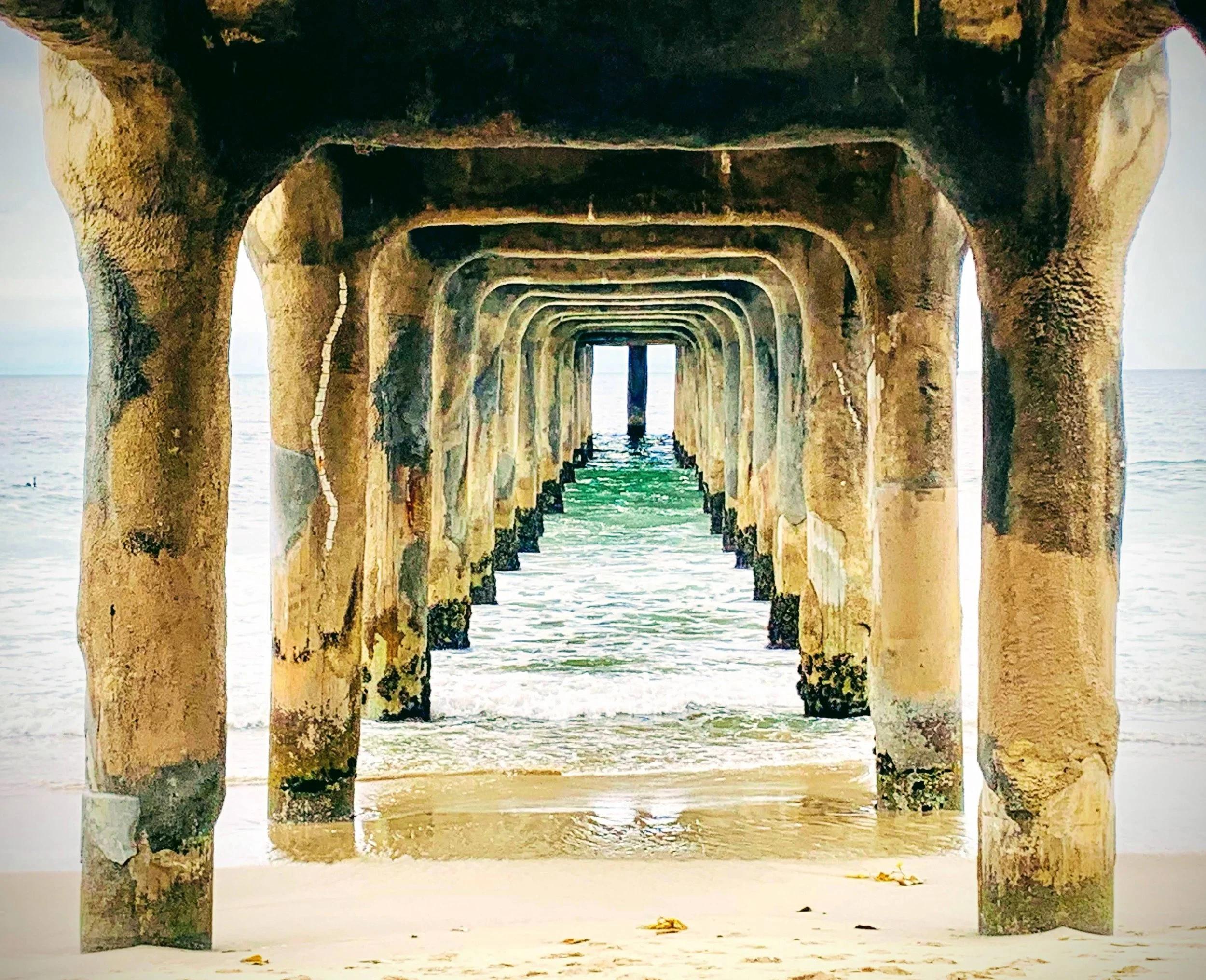 A view of a pier extending into the ocean, supported by concrete pillars, with water underneath and a sandy beach at the bottom.