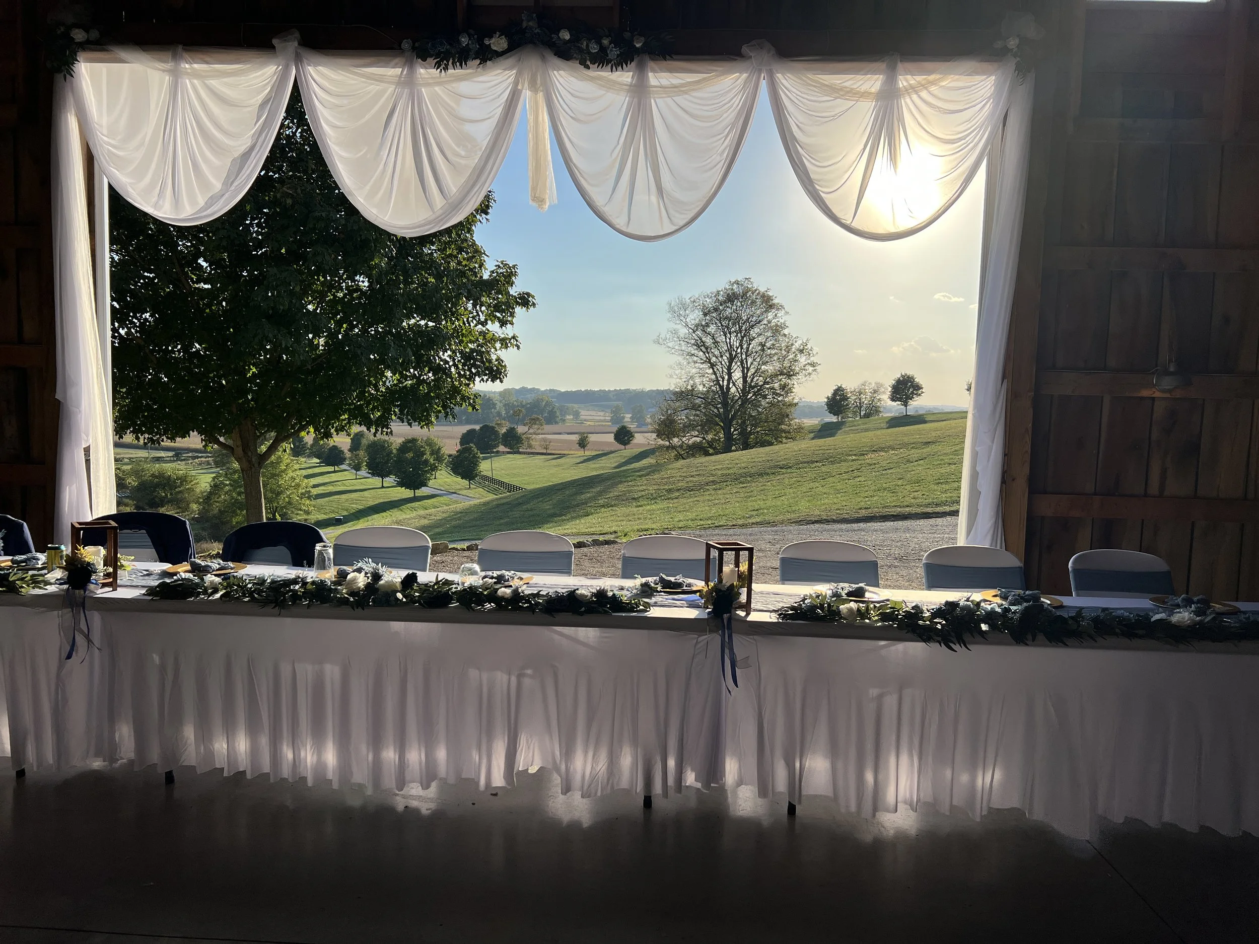 A decorated indoor banquet table set up with white tablecloth, floral arrangements, and lanterns, facing large open windows showing a scenic landscape of green rolling hills, trees, and a bright blue sky.
