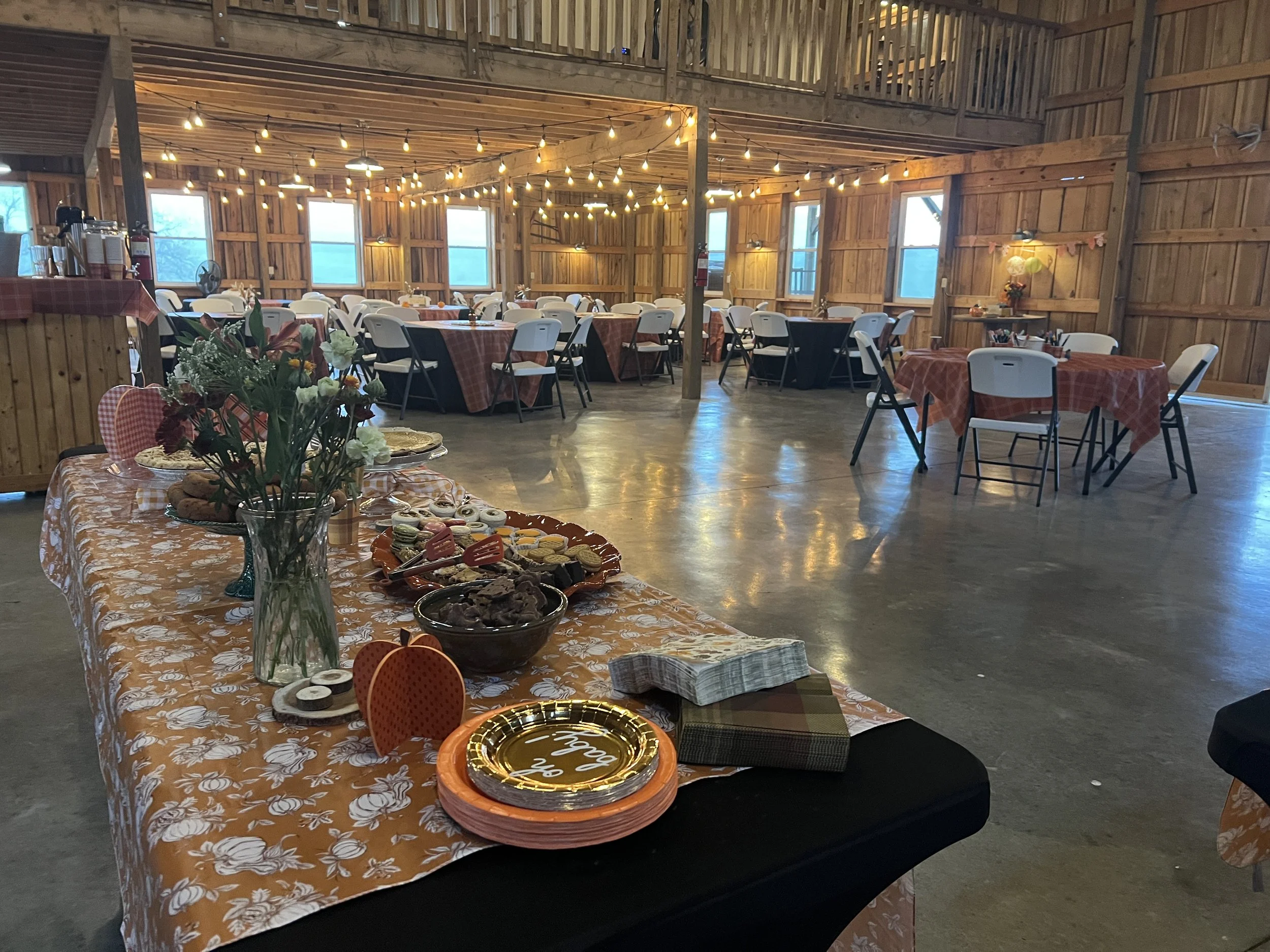 Interior of a rustic barn decorated for a celebration, with round tables covered in red checkered tablecloths, string lights hanging from the ceiling, a table in the foreground with snacks and cookies, and a flower arrangement in a glass vase.