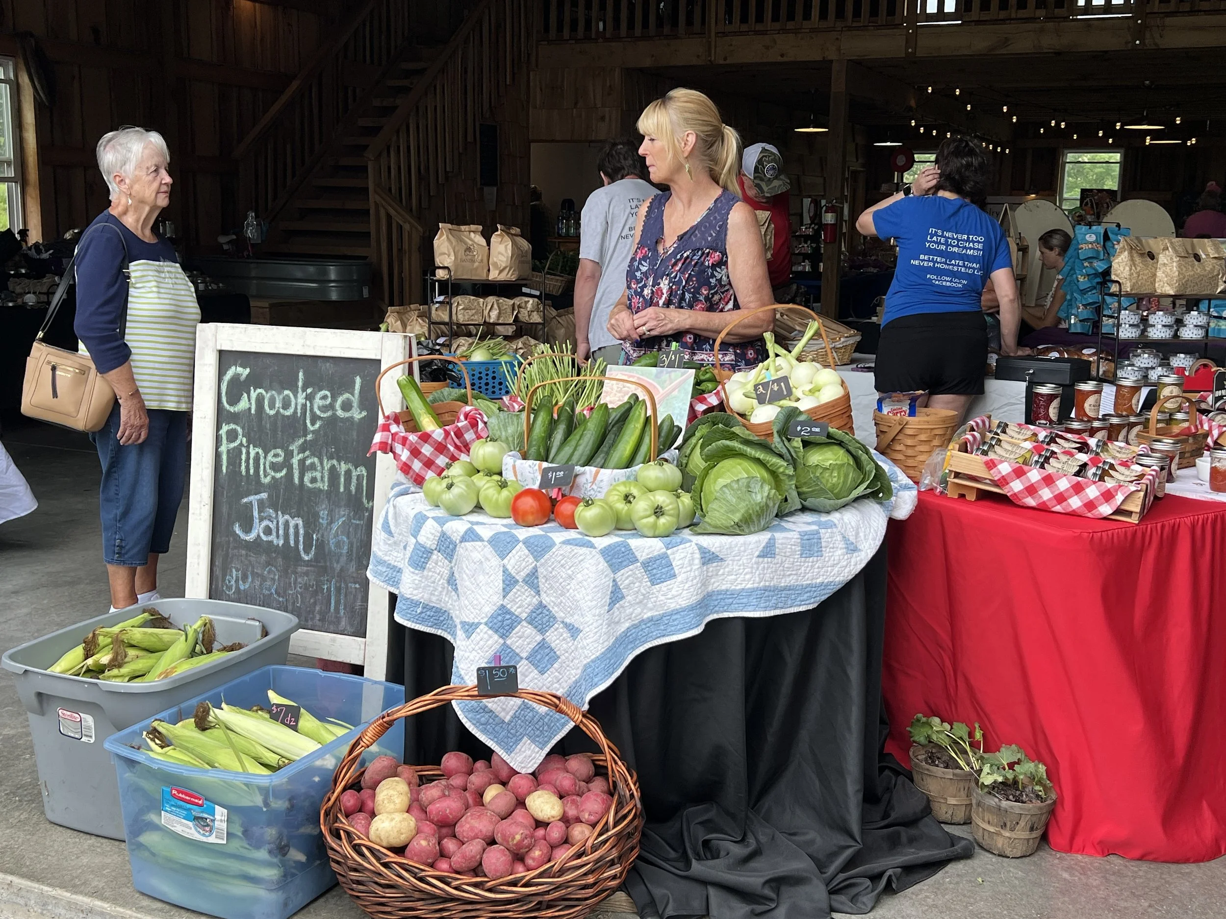 Women and a man at a farmers market stall with fresh vegetables including tomatoes, cucumbers, lettuce, and potatoes, with a blackboard sign advertising crookneck and pane tomato jam.