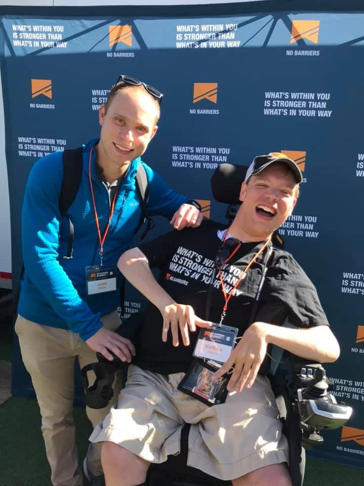 PHOTO: Matthew, in his wheelchair in a black shirt black hat and khaki shorts smiling. Standing next to him is his brother, Jason. He is smiling in a blue shirt and khaki pants They are having their picture taken in front of a blue backdrop that reads No Barriers.