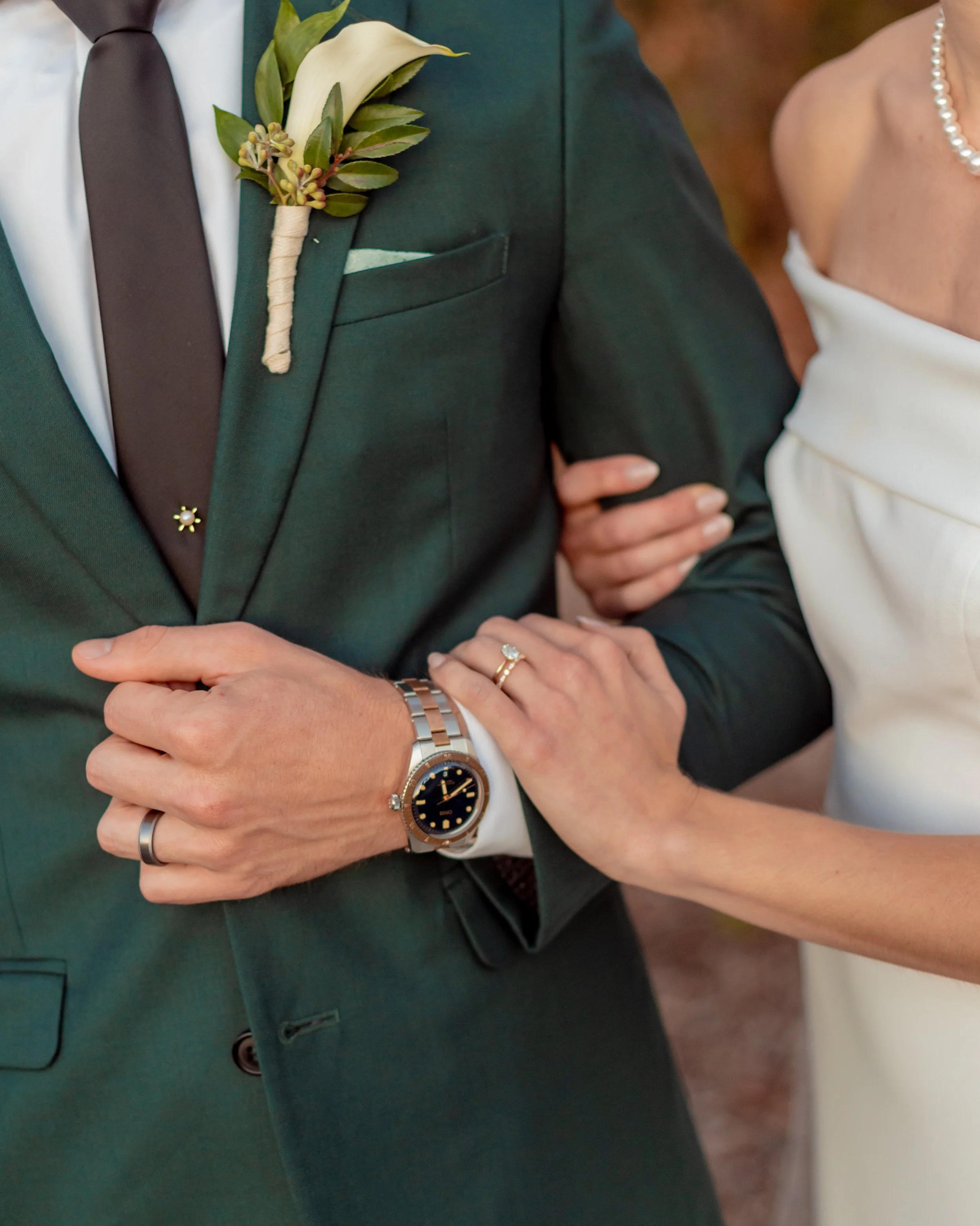  A close-up photo of a bride holding her groom’s arm. His hand is grasping the first button of his suit, creating a loop for her to hold. Her right loosely holds his bicep, while her left hand hold his wrist right over his black and silver watch. Bot
