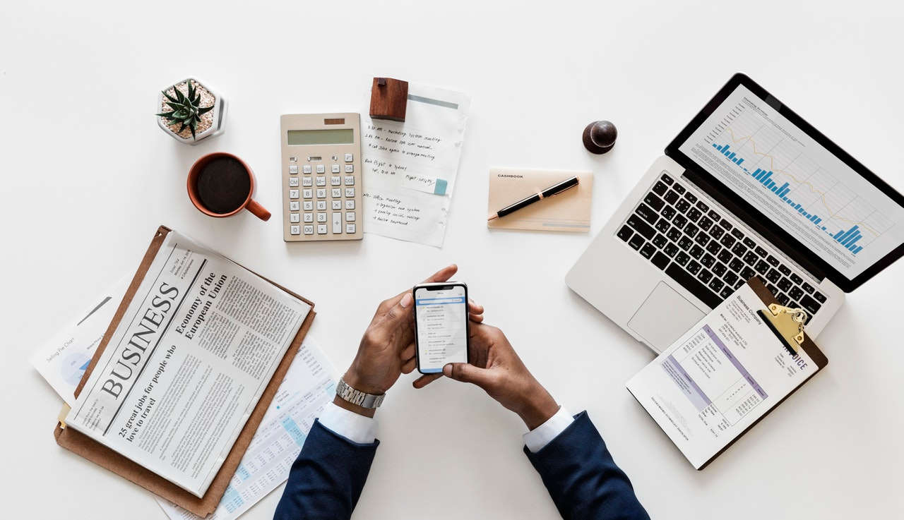 man in suit looking through phone over desk with laptop newspaper and calculator.jpeg