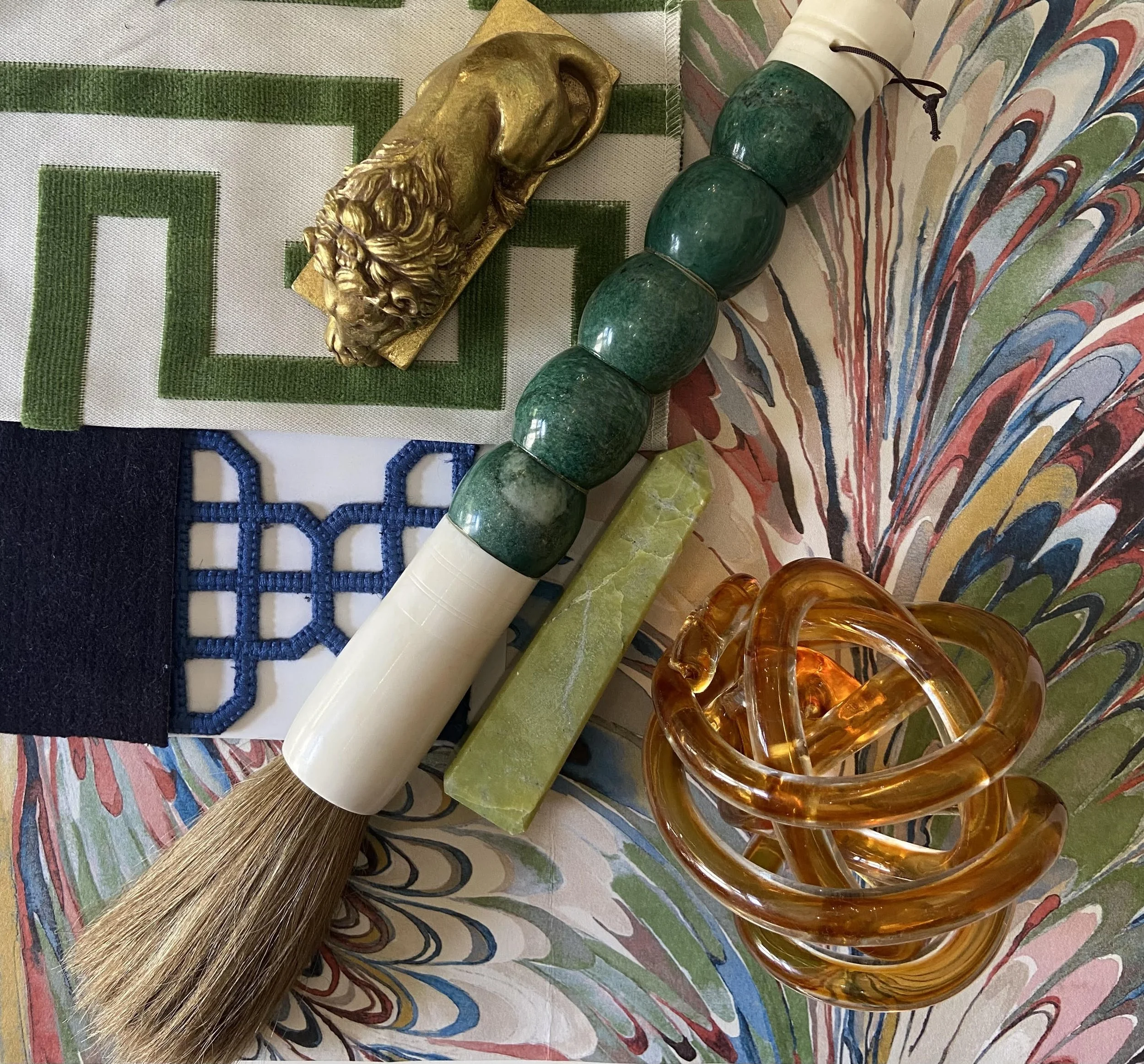 Collection of decorative objects including a golden lion figurine, a green bead necklace, a soap bar, an amber glass sculpture, fabric swatches, and a colorful marbled paper background.