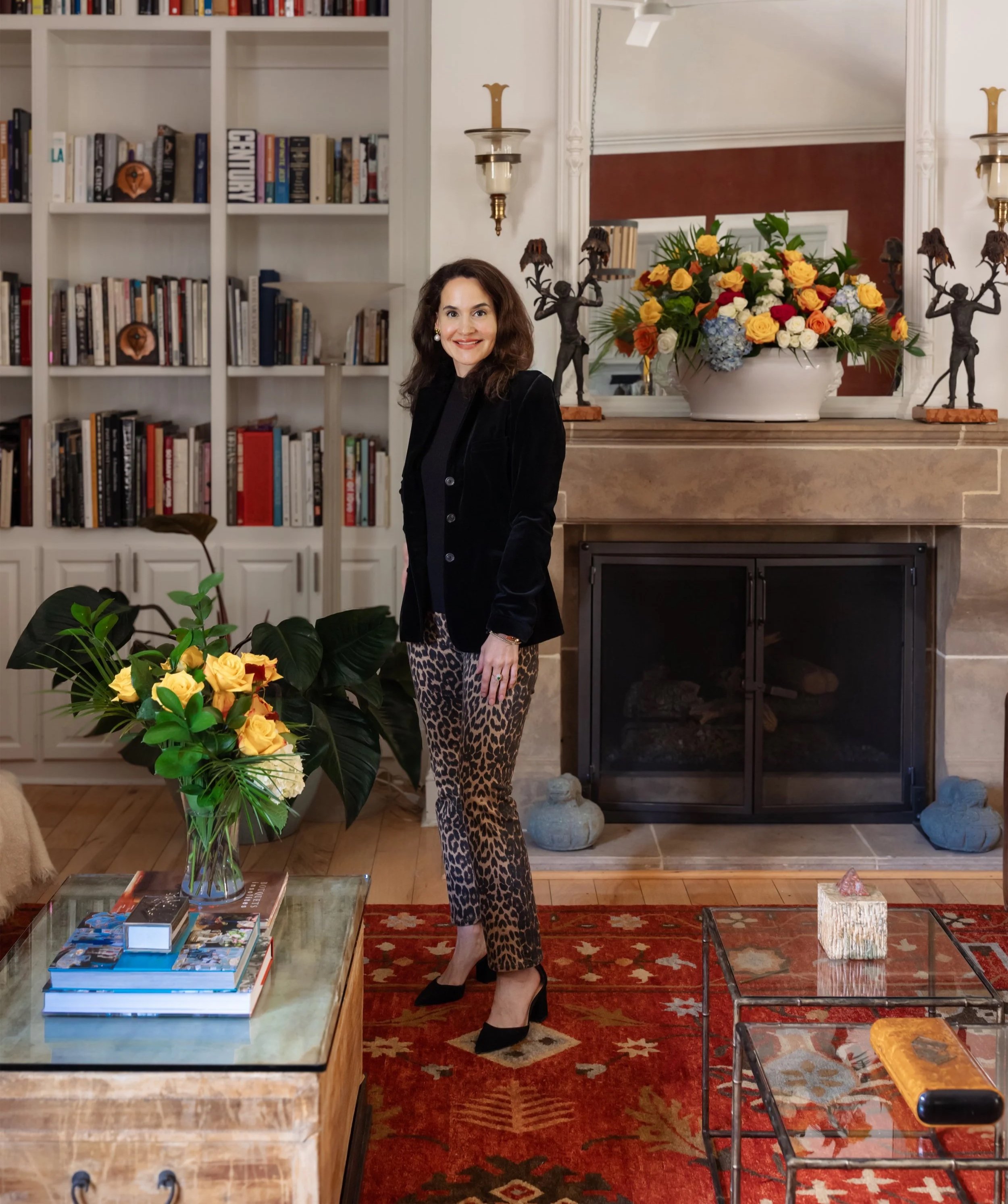 A woman standing in a living room with a fireplace, bookshelves, and decorative items, smiling at the camera.