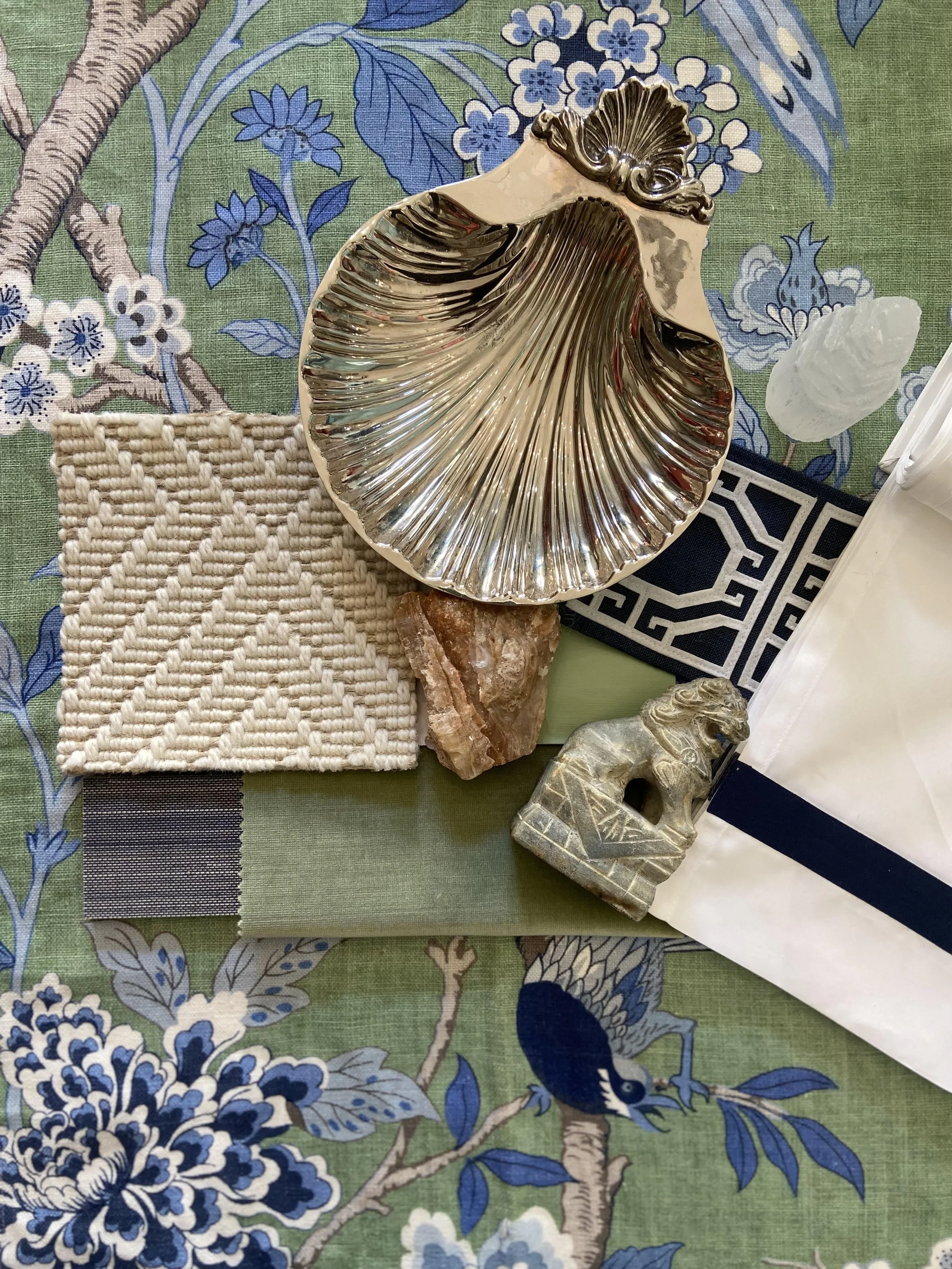 Decorative items arranged on a floral tablecloth, including a large metallic shell-shaped dish, a small woven textile, a piece of rough stone, a carved stone lion figure, and a container with a white and blue geometric design.