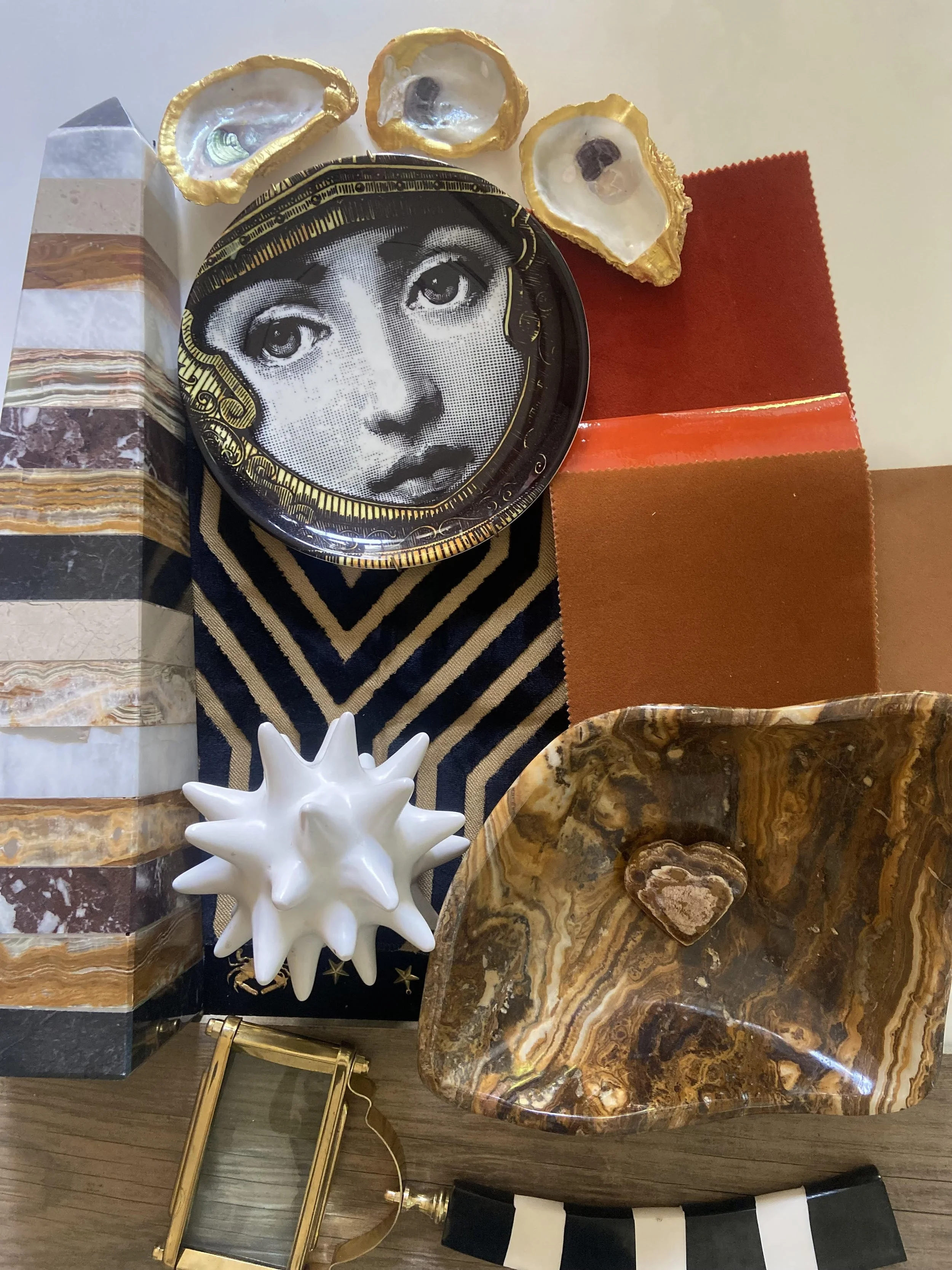 A collection of decorative objects including a black plate with a vintage woman's face, three oyster shells, a tall striped box, a white spiky ceramic sculpture, a patterned stone tray with a small heart-shaped rock, fabric swatches, a black and white striped item, and a small gold-colored metal frame.