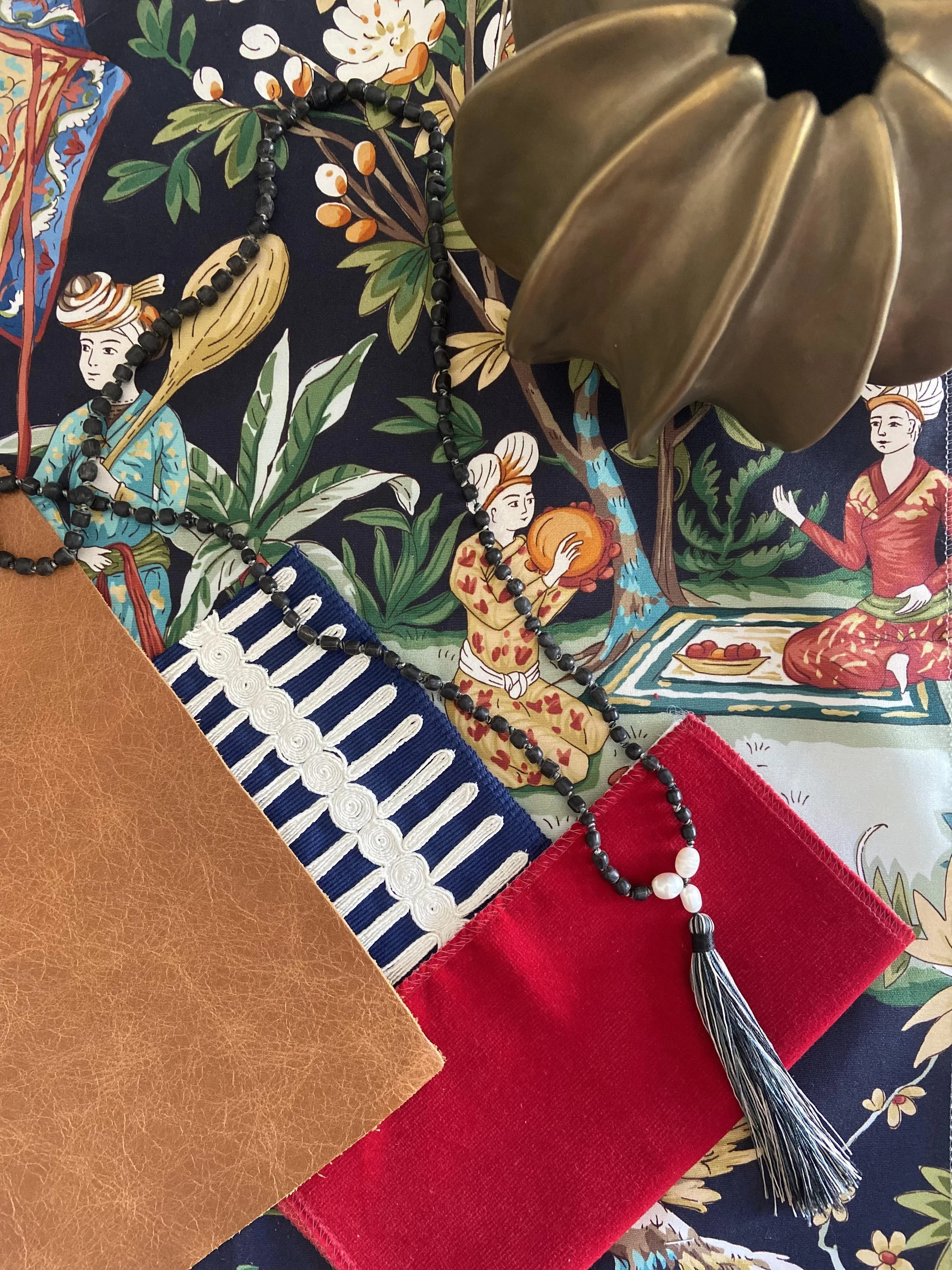 A table setting with decorative fabric and accessories, including a beaded necklace with a tassel, a piece of brown faux leather fabric, and a red cloth, with a background fabric featuring a colorful floral and human figure pattern and a brass leaf ornament.