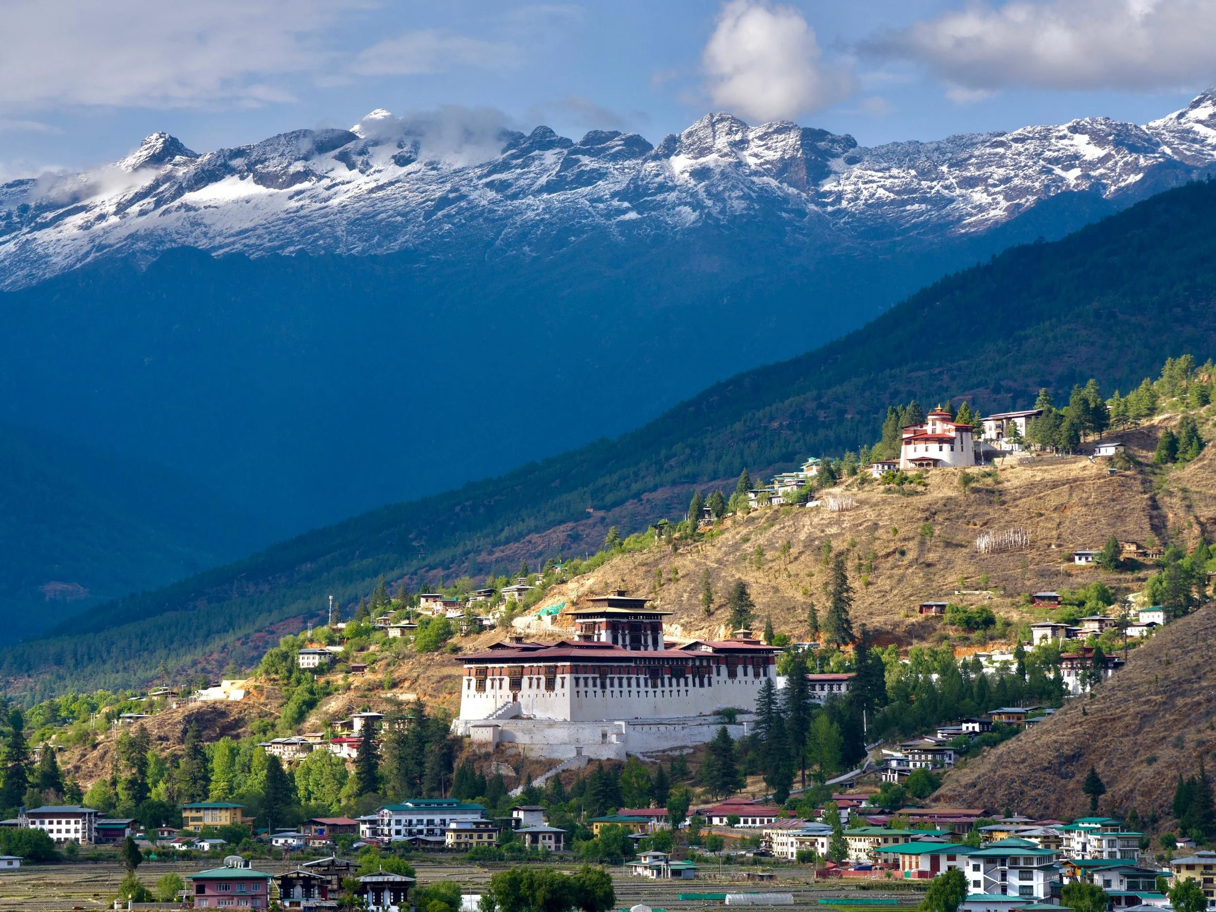 A large white monastery with red windows sits on a hillside against a background of snow-capped mountains and scattered buildings.
