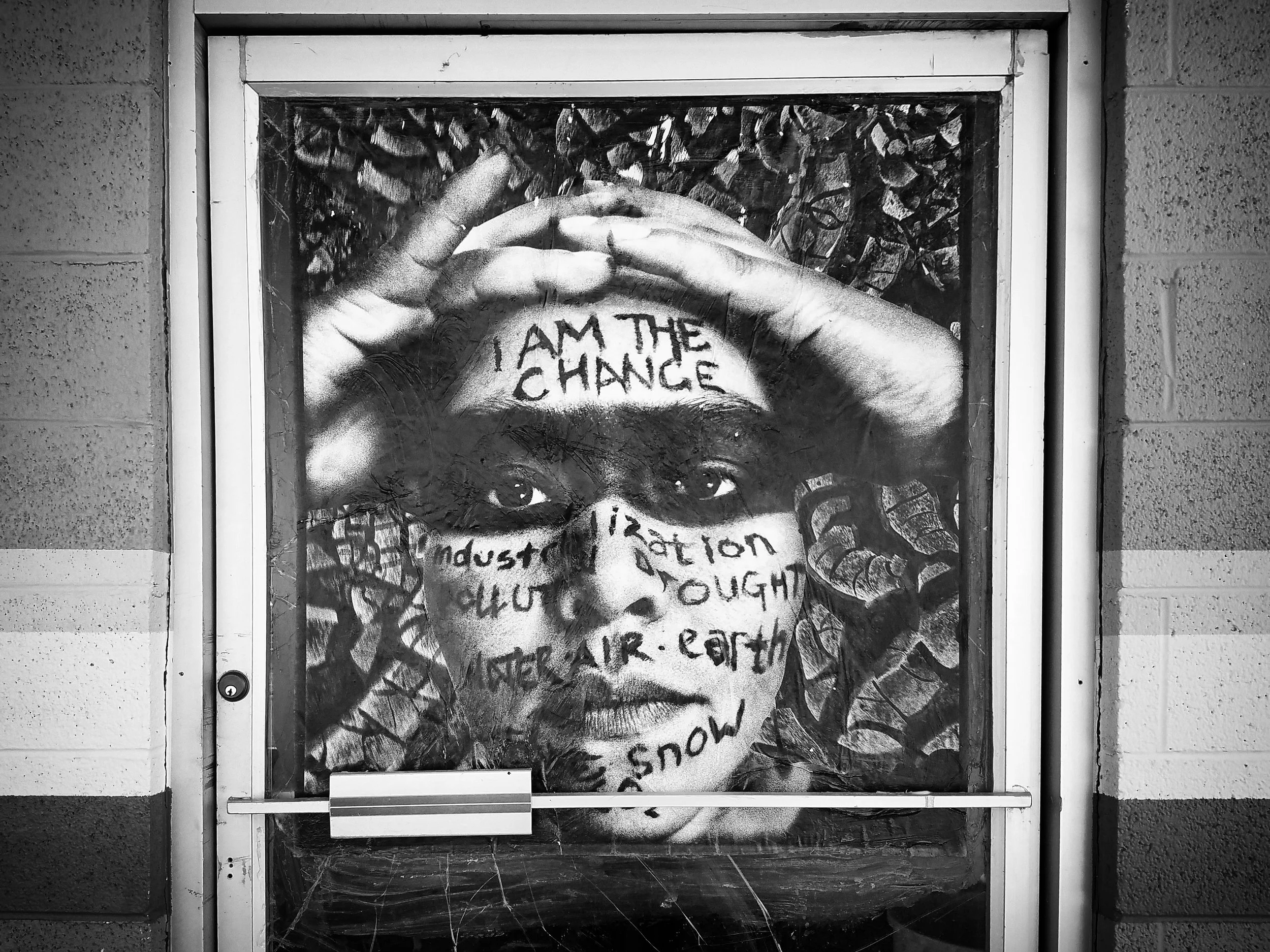 Street art of a woman's face painted on a glass door, with the phrase 'I AM THE CHANGE' written across her forehead. The artwork includes eye details and text overlay.