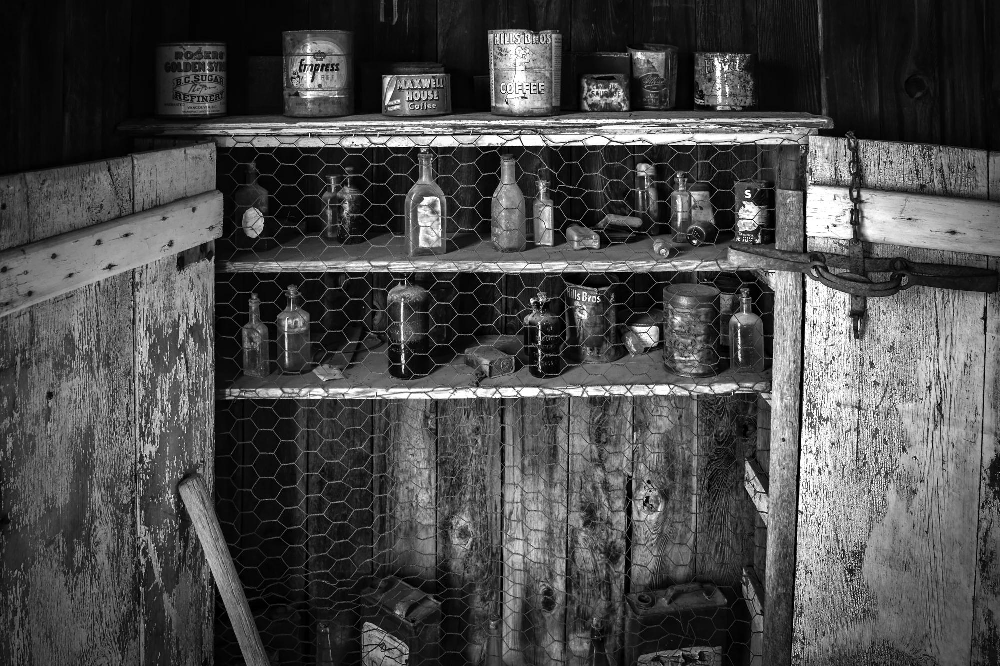 Black and white photo of a rustic wooden cabinet with a wire mesh door containing old glass bottles. The top shelf holds vintage metal containers labeled with old-fashioned brand names.