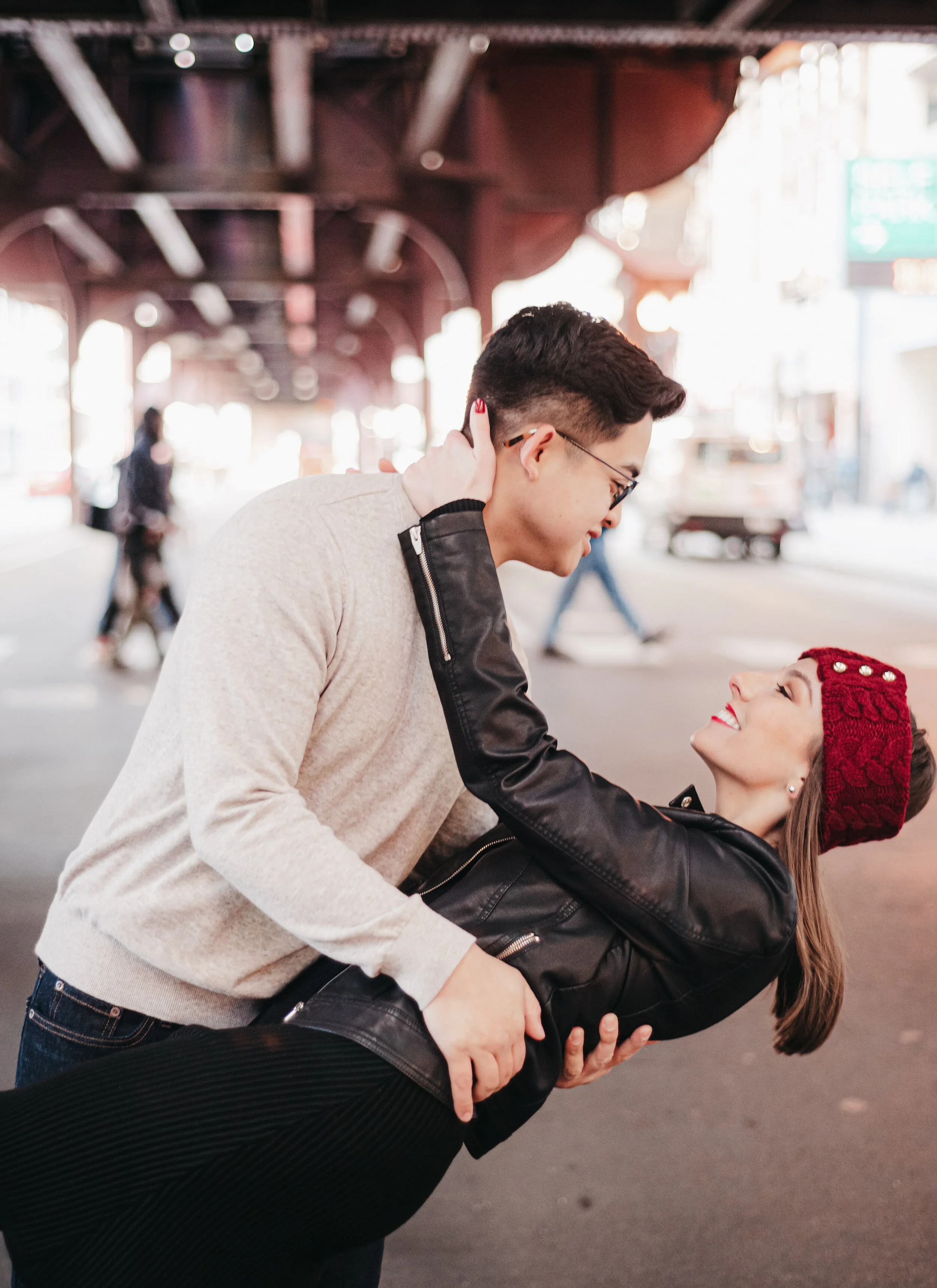 A young couple is dancing and smiling on a city street under an overpass, with blurred pedestrians walking in the background.