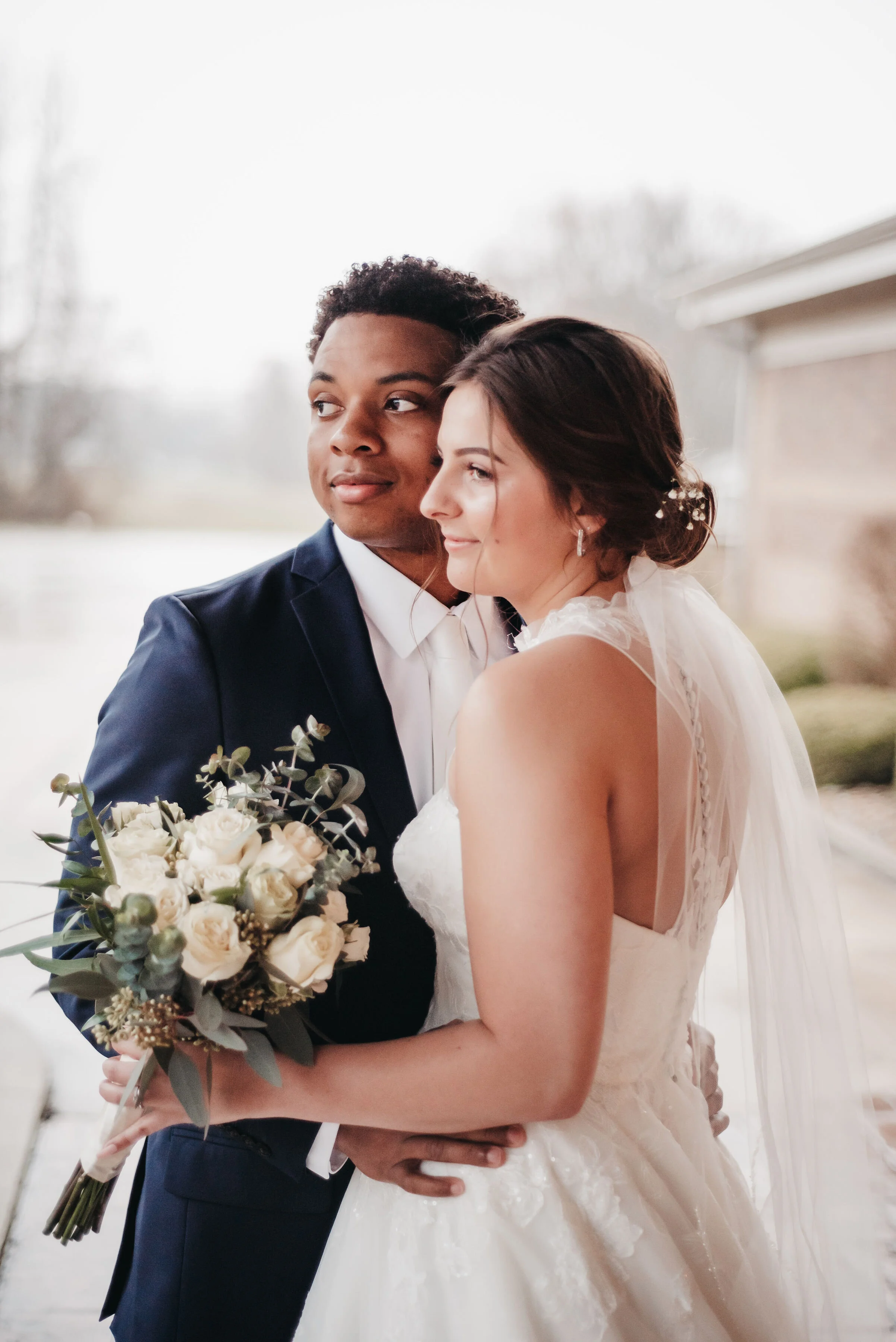 A newlywed couple dressed in wedding attire standing close together outside, with the bride holding a bouquet of white roses and greenery, and the groom in a navy blue suit. The background shows a slightly overcast outdoor setting.