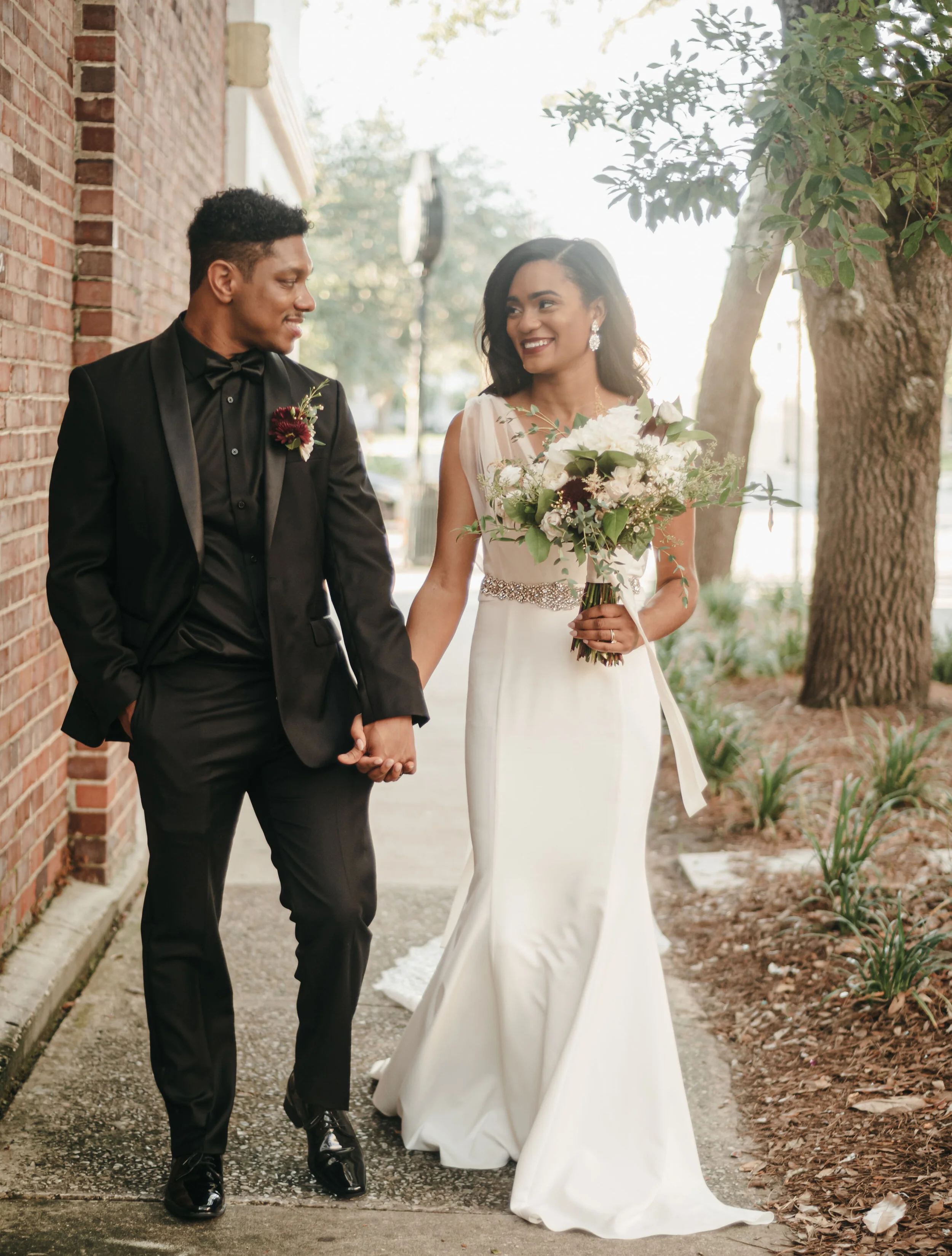 A bride and groom walking hand in hand outdoors, smiling at each other, with the bride holding a bouquet of flowers.