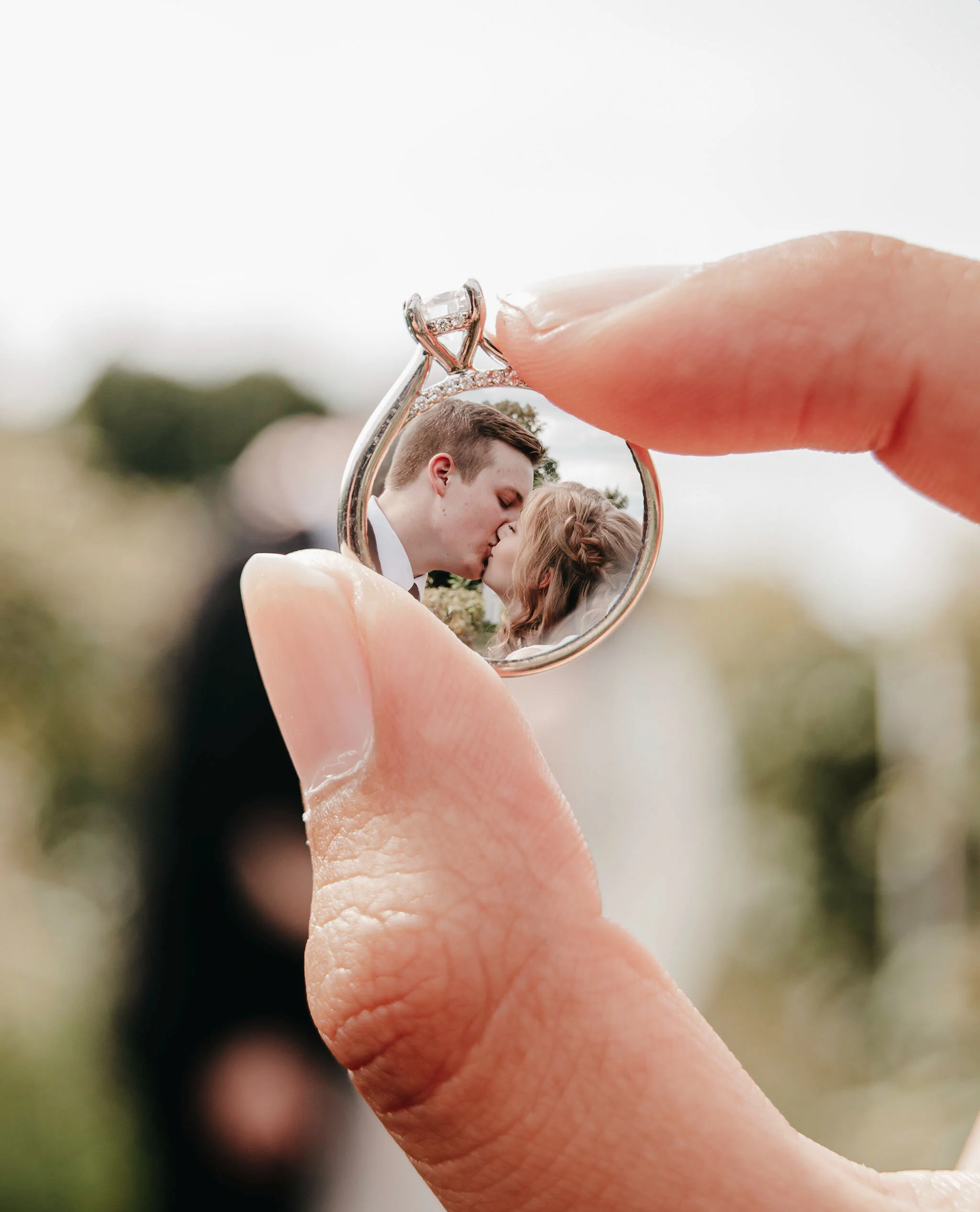 Close-up of a hand holding a small mirror reflecting a bride and groom kissing.