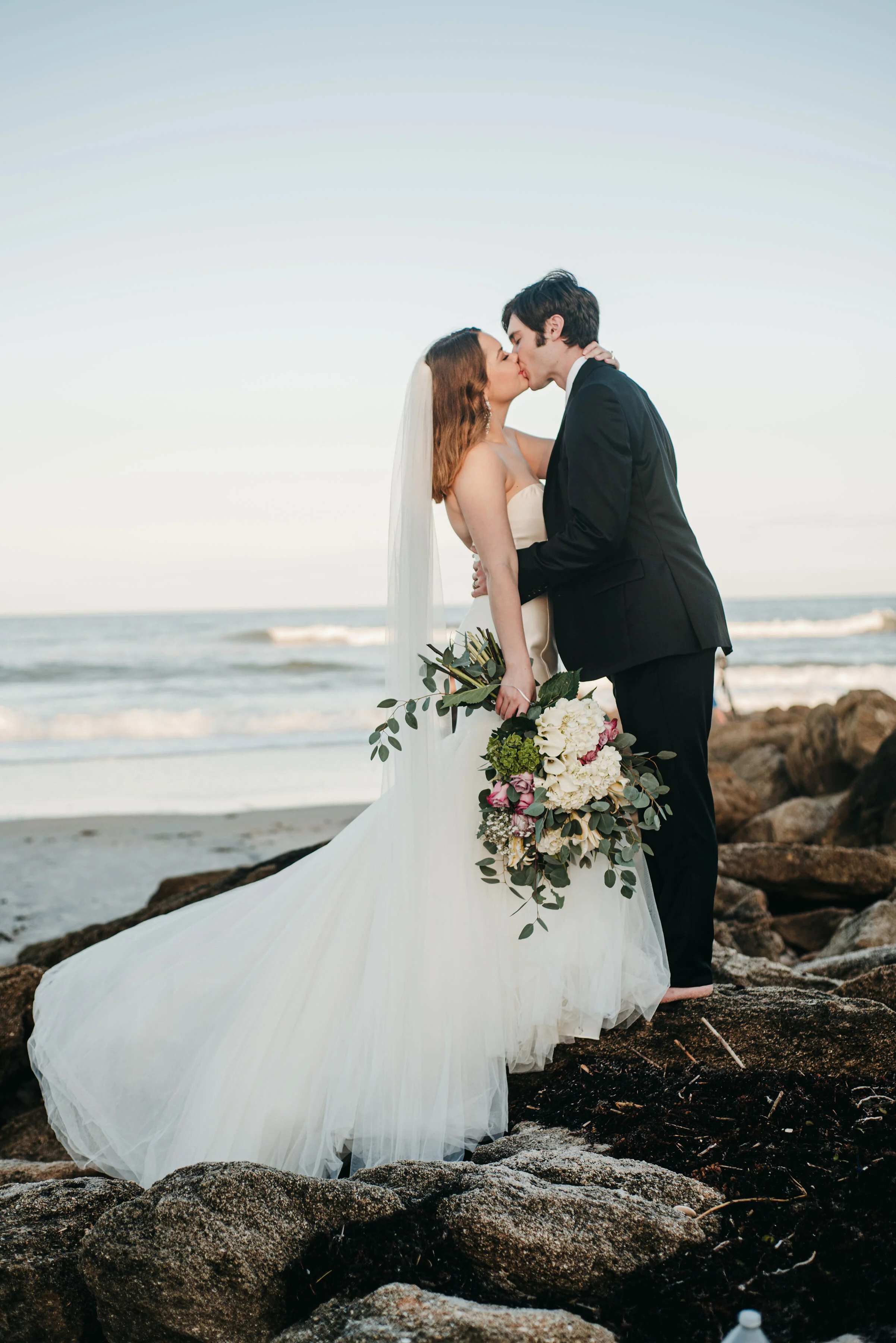A bride and groom share a kiss on a rocky beach at sunset, with the bride holding a bouquet of white and purple flowers.