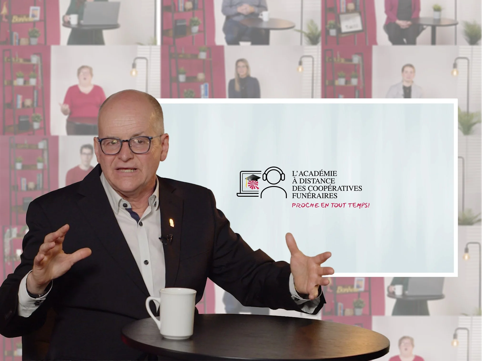 Un homme en costume noir parle devant un écran présentant un logo et un texte en français. L'écran affiche le texte : 'L'Académie à distance des coopératives funéraires proche en tout temps!'.  C'est une vidéo de formation tournée au plateau.