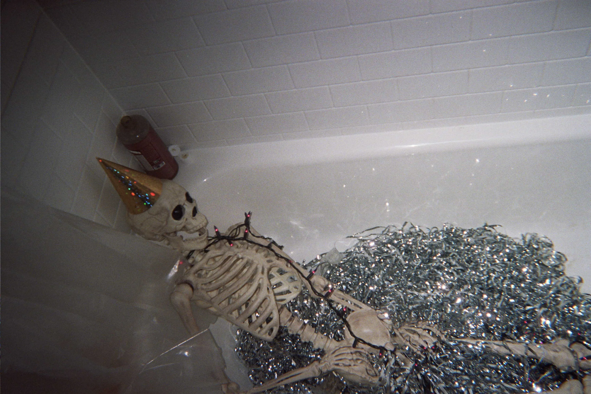 Skeleton wearing a green party hat with Christmas lights, lying in a bathtub filled with silver tinsel, with a bottle of wine and a white object nearby.