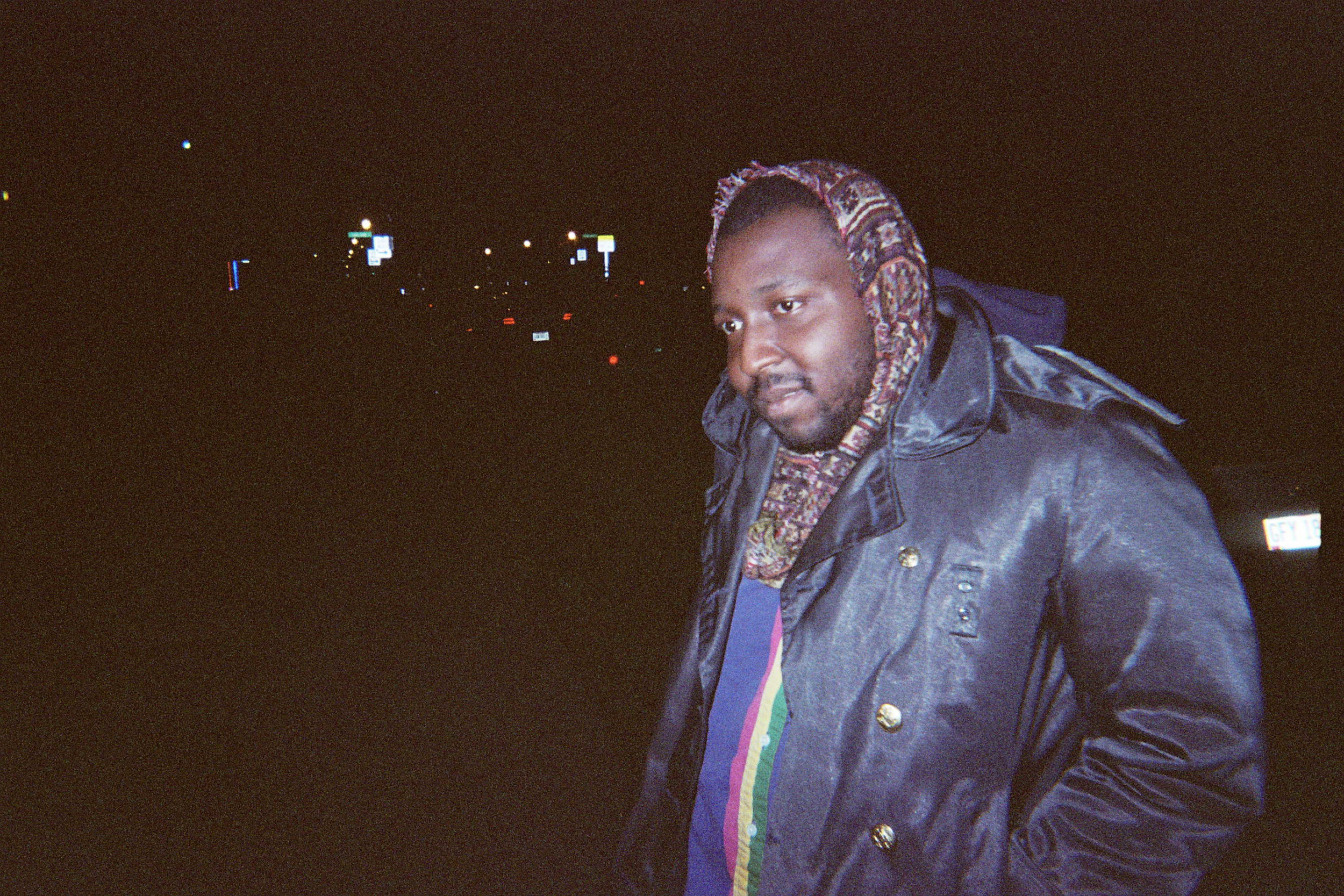 A man standing outdoors at night, wearing a dark leather jacket, a colorful scarf, and a patterned hood. City lights and road signs are visible in the background.
