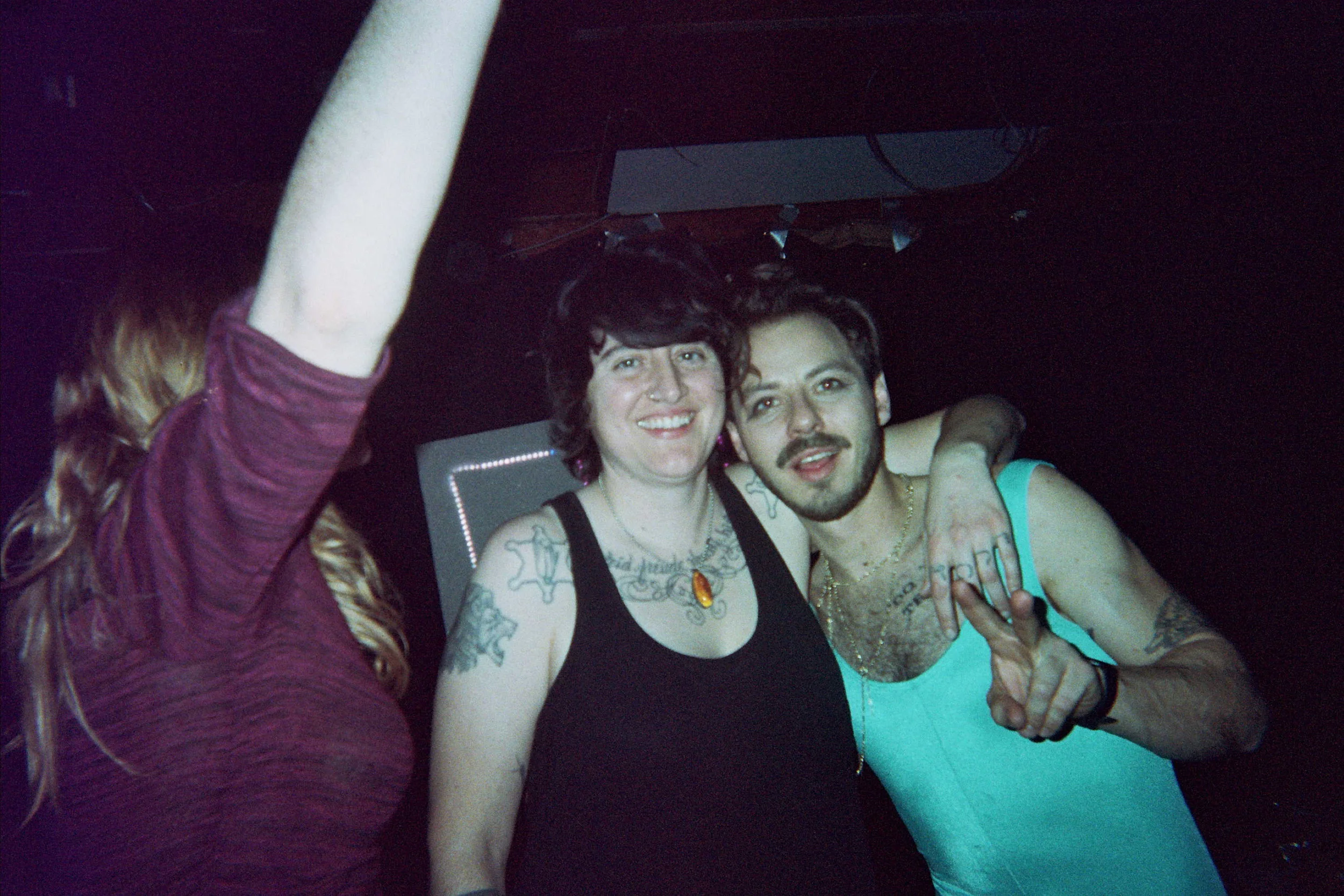 Two people smiling at a party or nightclub, with their arms around each other, and a third person partially visible on the left raising an arm.