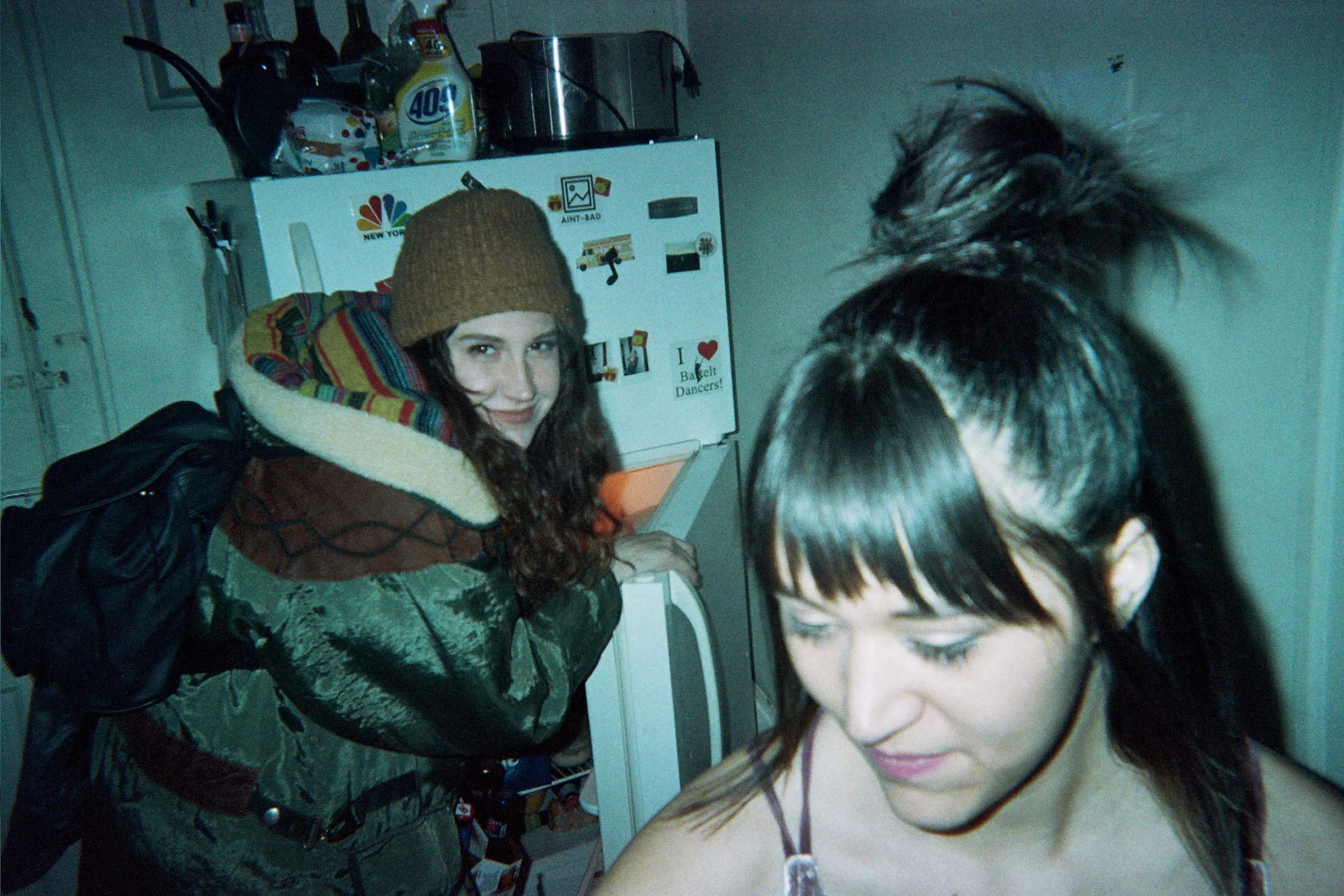 Two young women in a kitchen, one is wearing a brown beanie hat, a brown and green jacket with a colorful scarf, and has long curly hair; the other has long dark straight hair with bangs, and has her hair styled in a spiky ponytail. They are standing