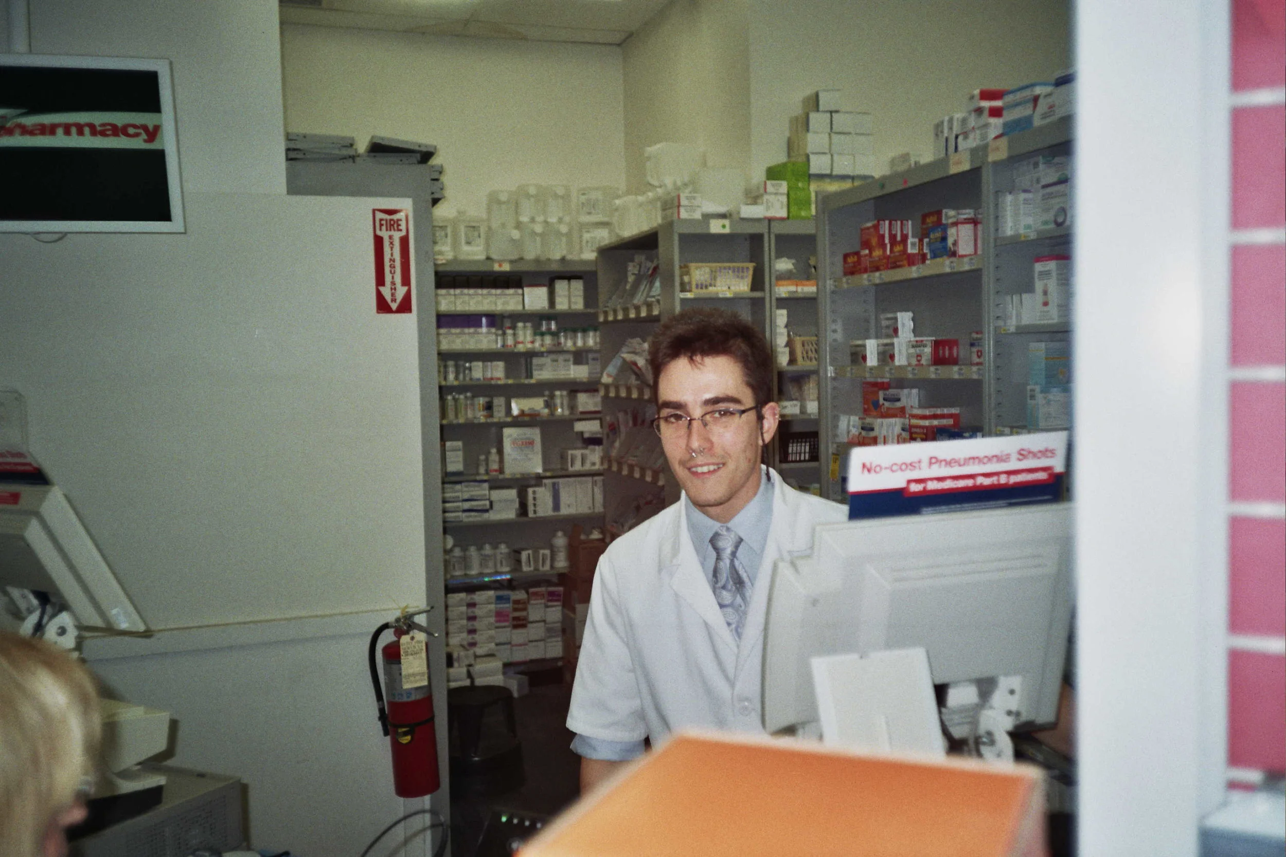 A young man with glasses and a mustache wearing a white lab coat, smiling at the camera inside a pharmacy with shelves of medications in the background.