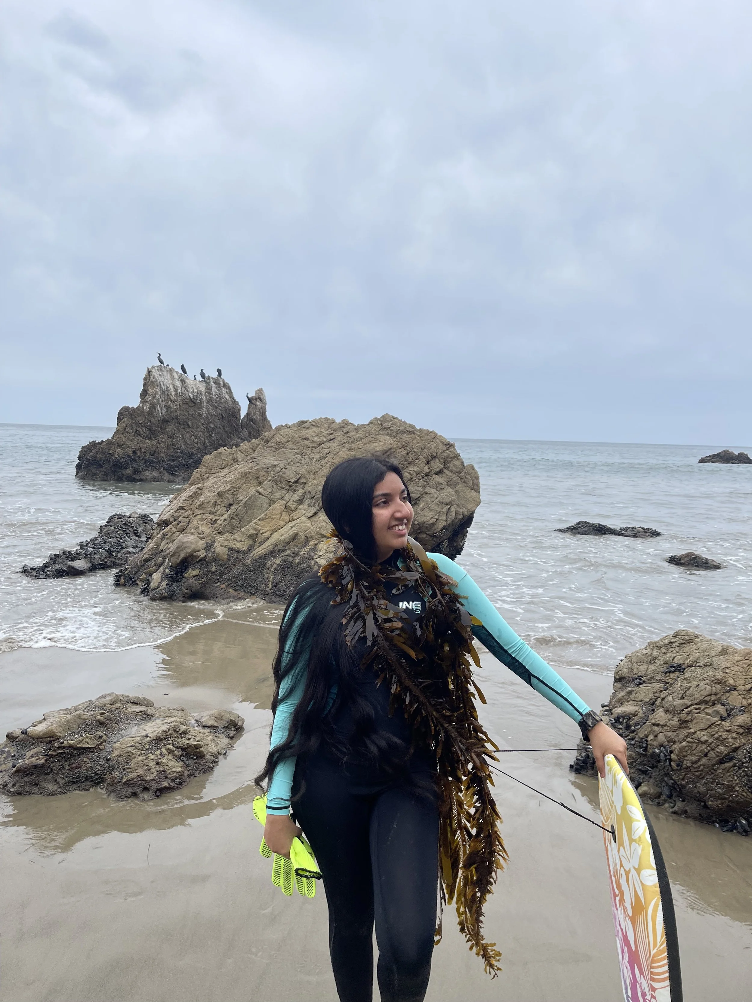 Gwenevere Persaud on a beach wearing a wetsuit and holding a bodyboard with kelp draped around their neck
