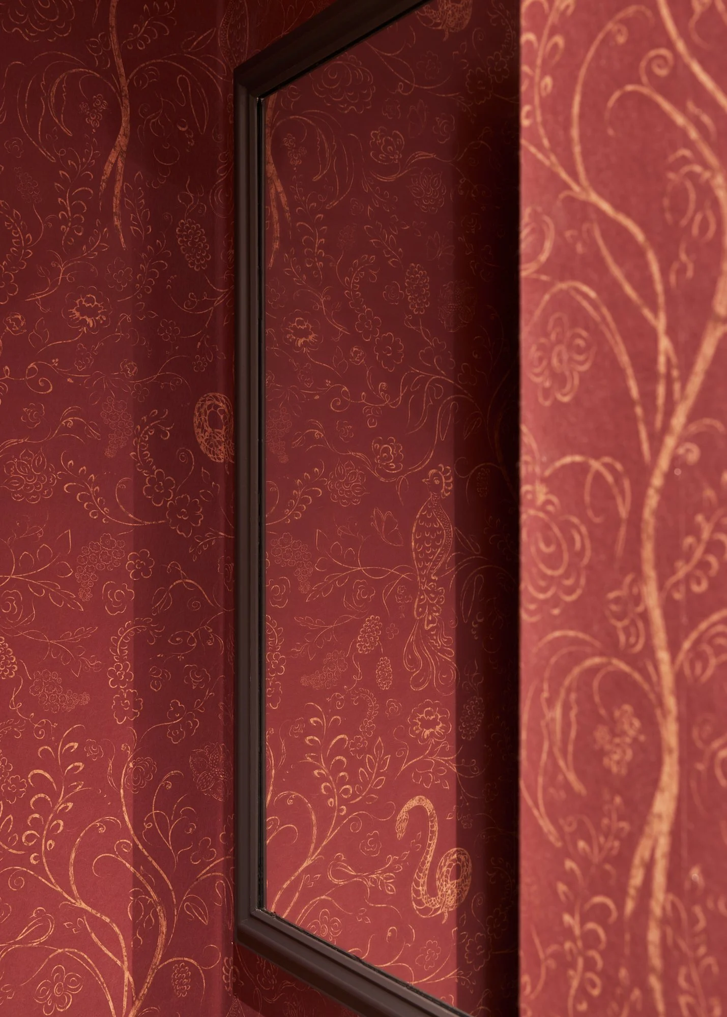 A wall with red wallpaper featuring gold floral and peacock patterns, and a rectangular mirror with a dark brown frame reflecting the wallpaper.