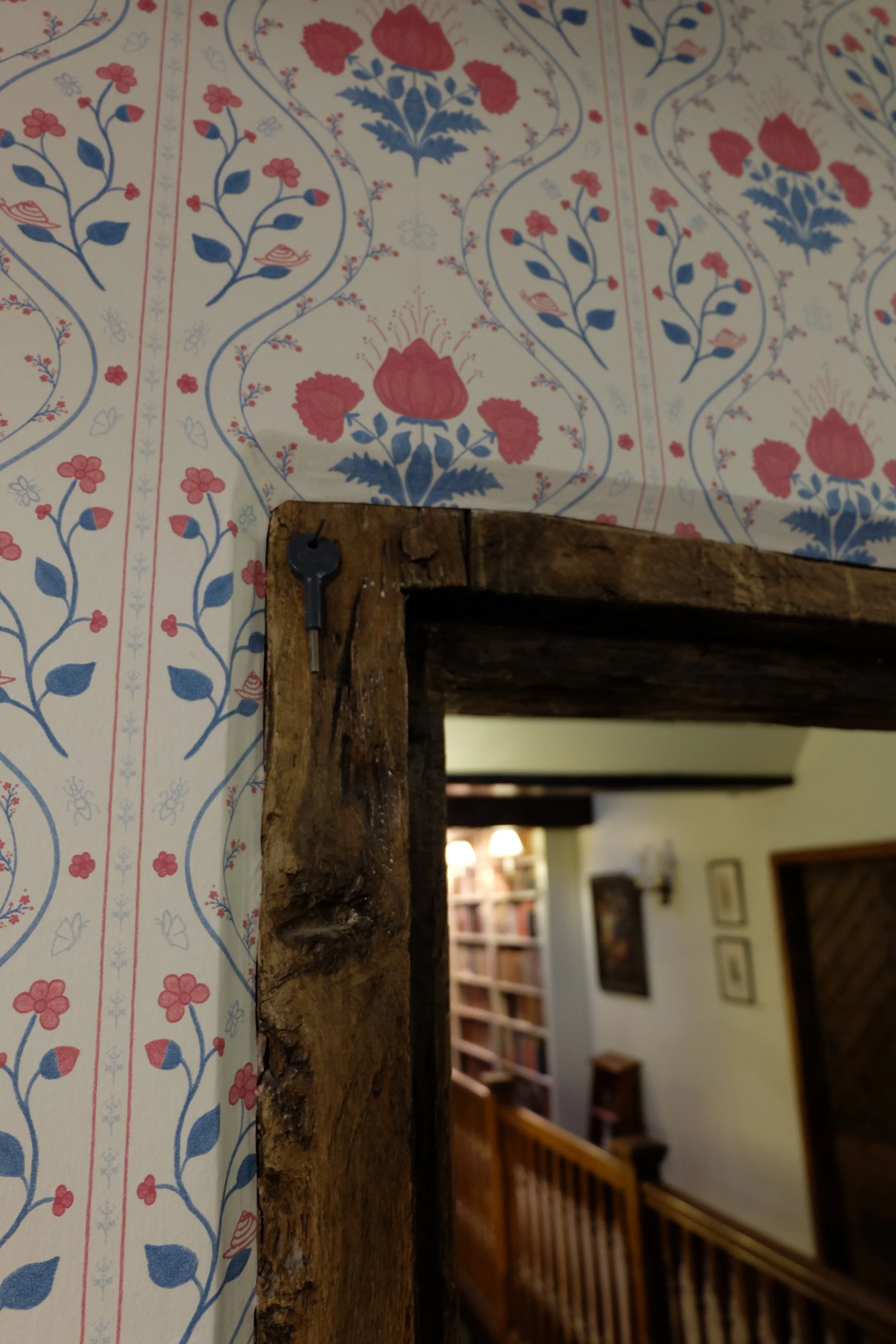 Close-up of a wooden door frame with a small key inserted into a lock, next to vintage floral wallpaper with red and blue flowers.