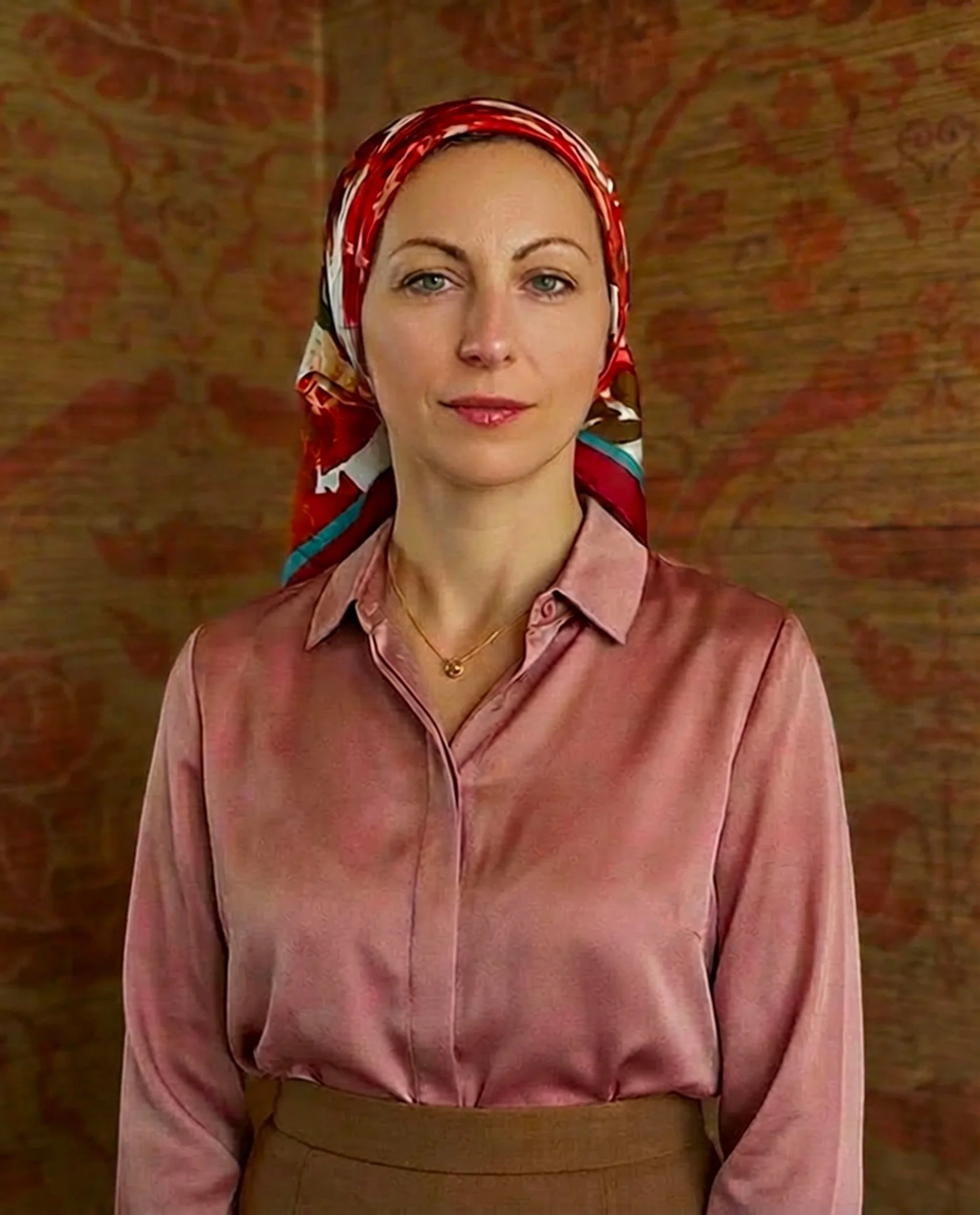 A woman with fair skin and blue eyes wearing a colorful headscarf, a pink satin blouse, and a gold necklace standing in front of a patterned background.