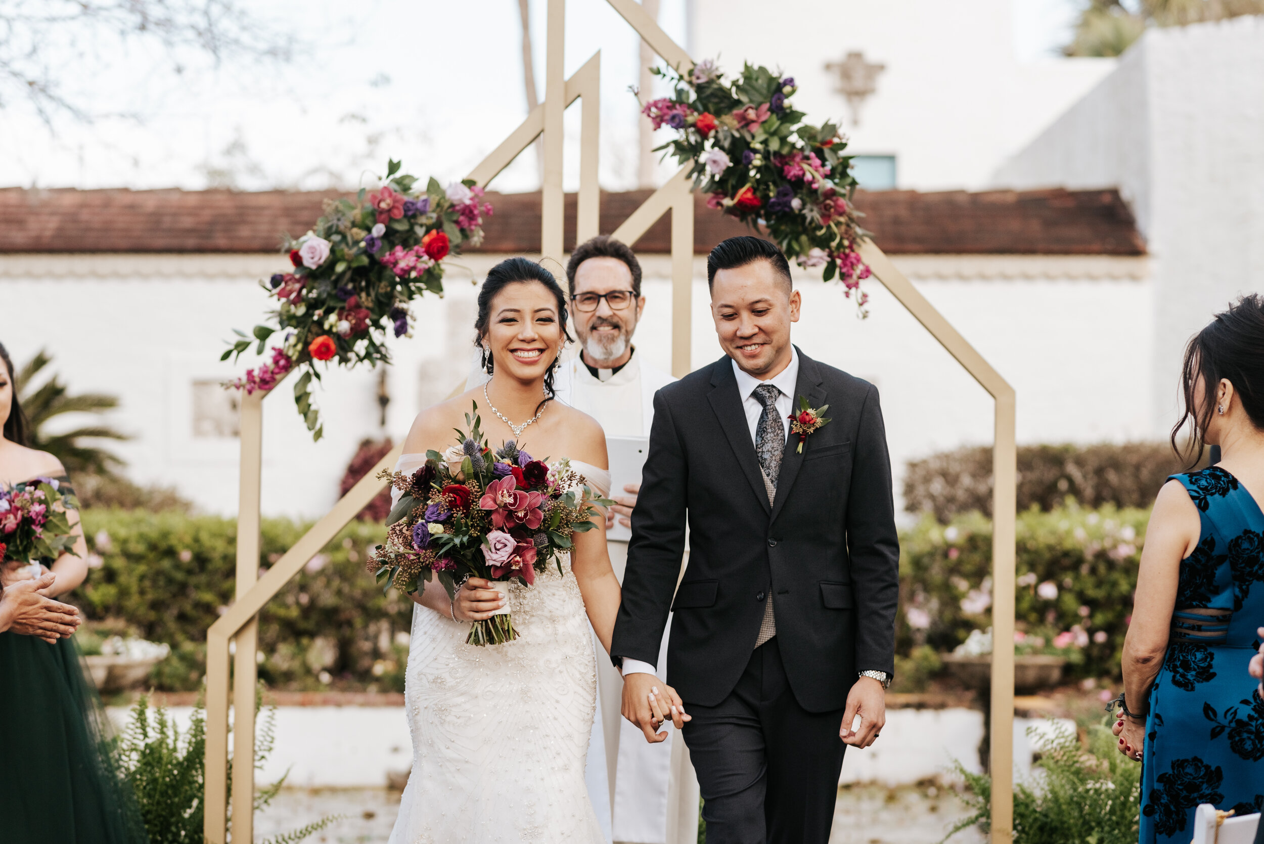 The bride and groom in front of their geometric arch decorated with jewel tone floral pieces (Copy) (Copy)