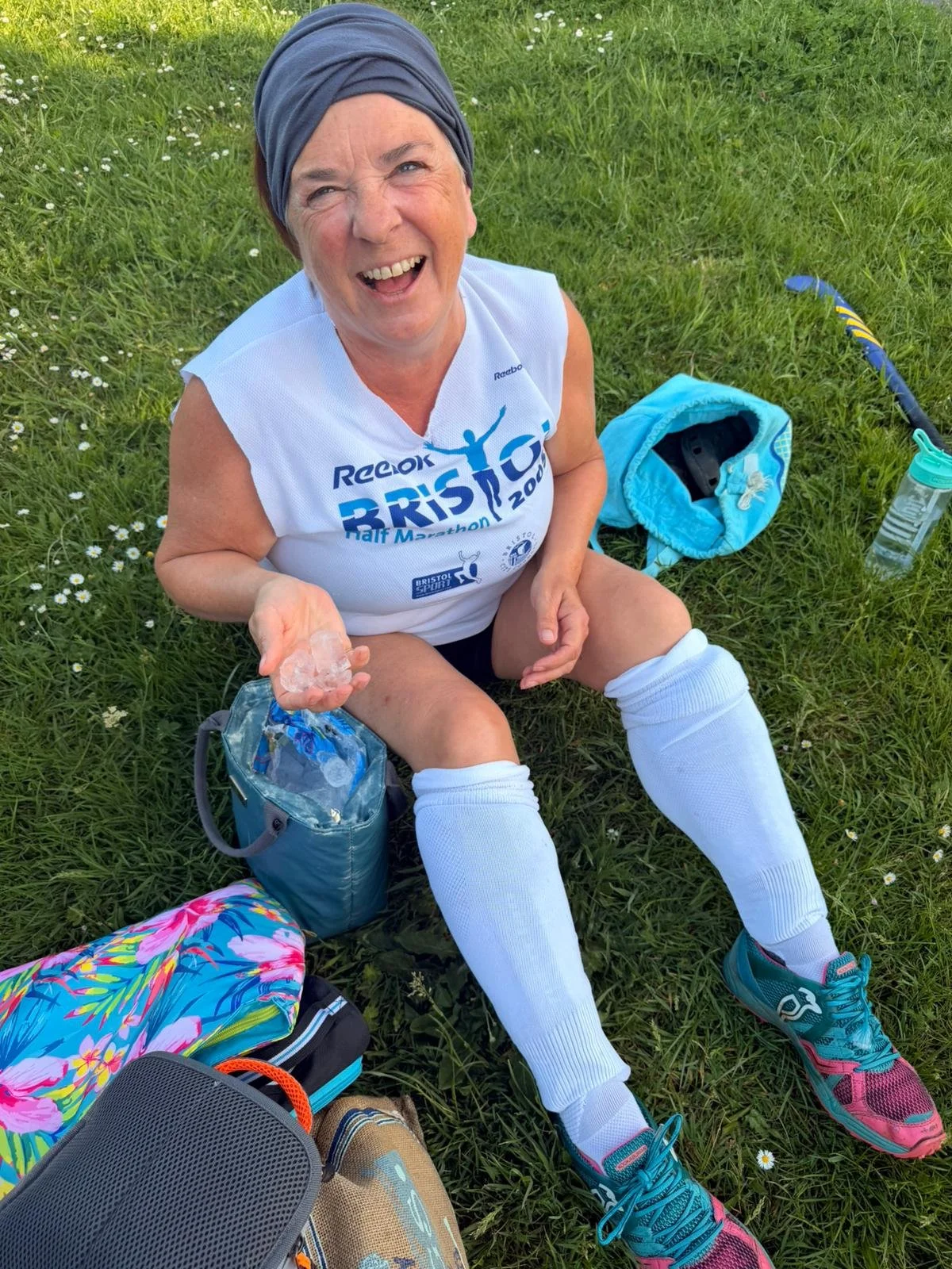 A woman sitting on grass after a race, holding ice and smiling. She wears a white athletic shirt, white knee-high socks, pink and blue running shoes, a headscarf, and shorts. There are backpacks and water bottles around her.