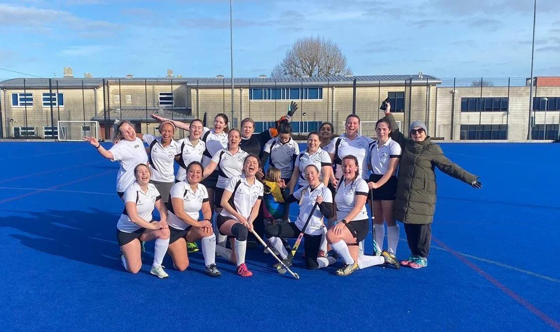 A group of young women in white sports uniforms on a blue hockey field, celebrating after a game with joyful expressions, some kneeling and others standing, surrounded by coaching staff and equipment.