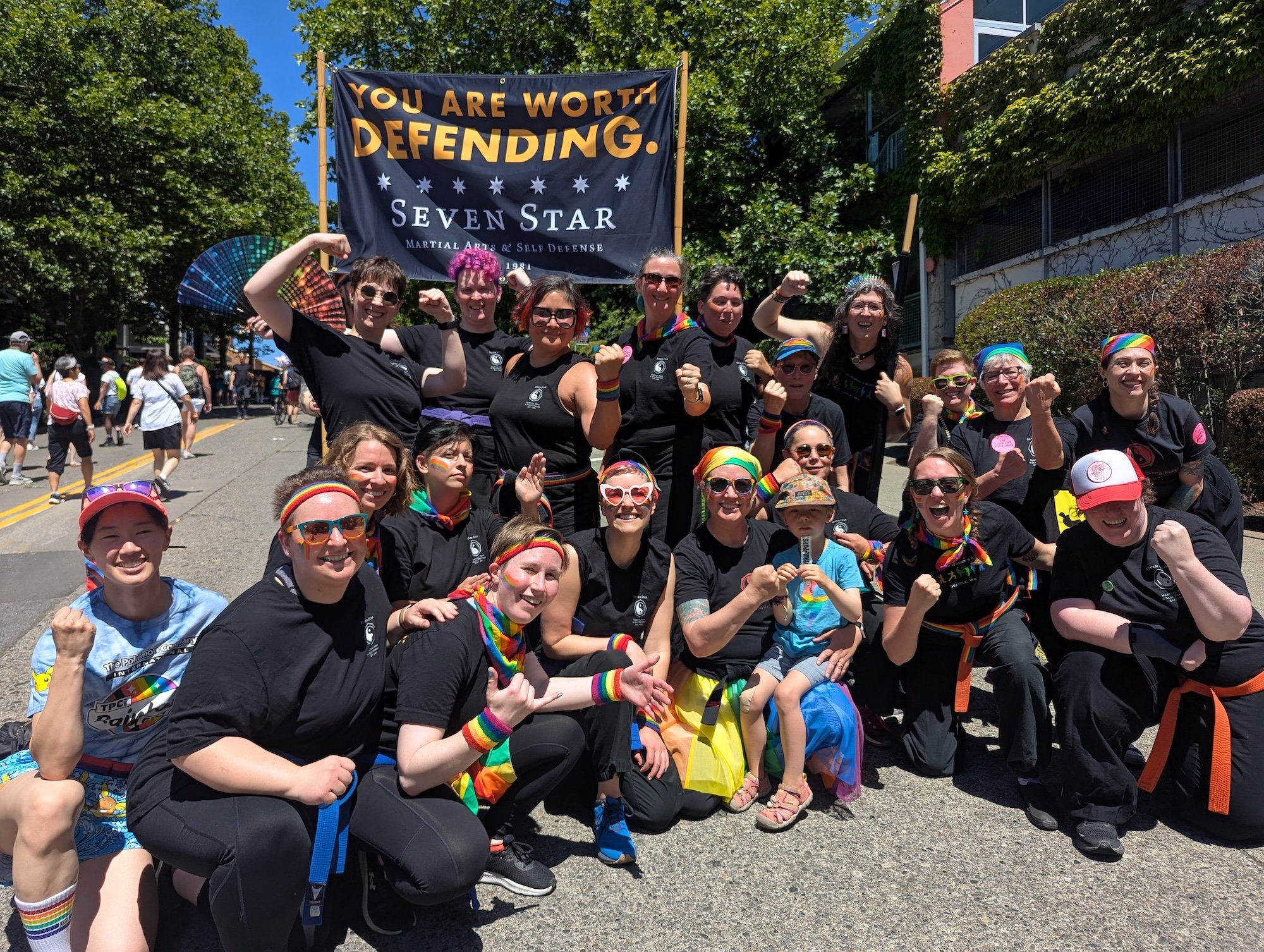 A group of trans, non-binary, and cis women martial arts are posed cheering at Seattle's Pride parade in front of a banner that says "Your Are Worth Defending- Seven Star"