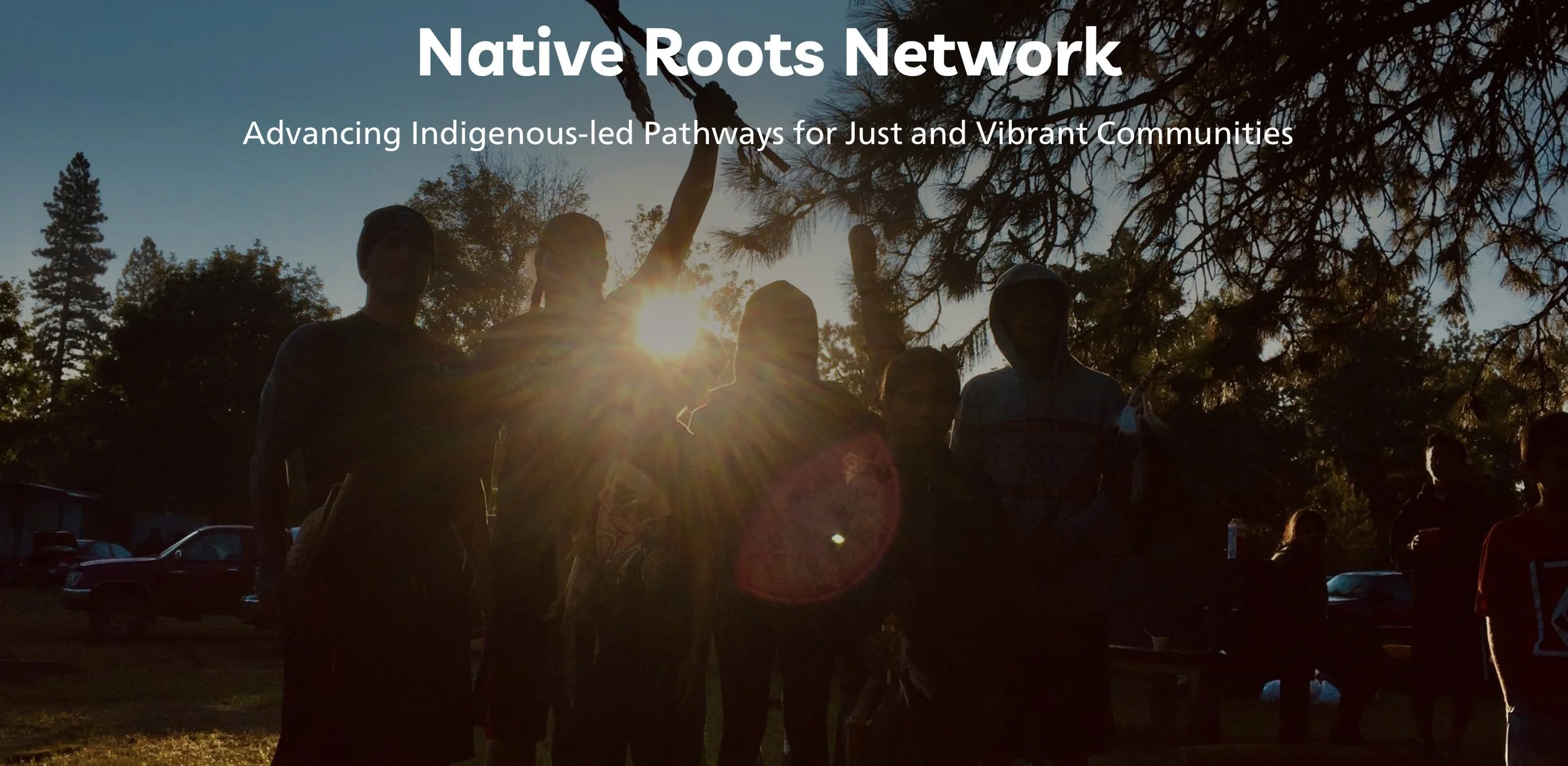 California Tribal people stand, backlit,  with the setting sun at their backs in a tree filled clearing. One individual holds a ceremonial staff high in the air.